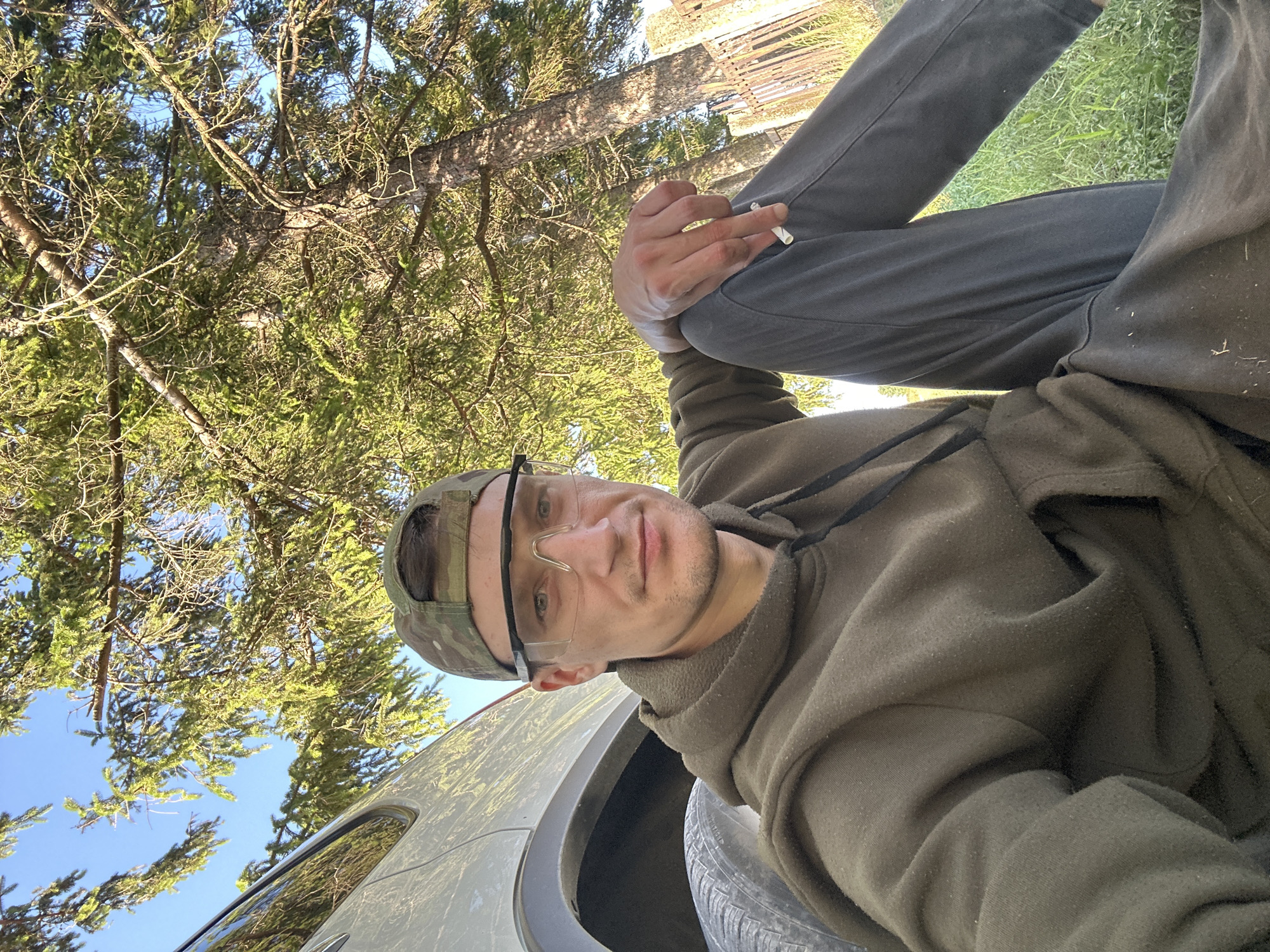 A young man wearing a camouflage baseball cap, safety glasses, and an olive-green hoodie sits on the grass next to a light-colored vehicle. He is holding a cigarette between his fingers. The background shows a large tree with green needles, a blue sky, and a wooden fence. The scene appears to be outdoors, likely in a rural or semi-rural area, possibly Makiv, Ukraine, given the context provided. The lighting suggests it is daytime, with sunlight filtering through the tree branches.