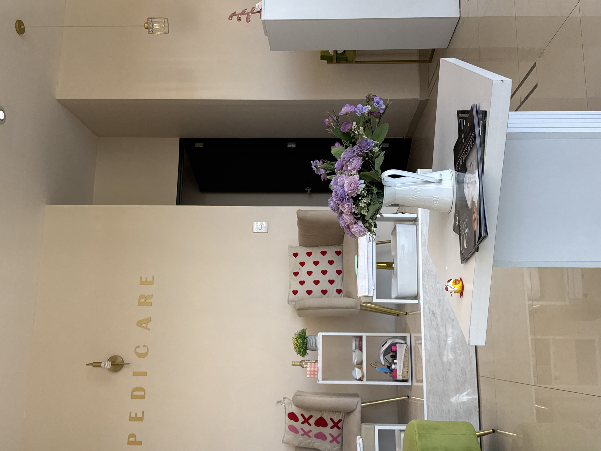 A brightly lit interior of a pedicure salon in Puebla, Mexico, shows a beige wall with vertical gold lettering spelling "PEDICARE" next to a vintage electrical fixture. Two beige armchairs with heart-patterned and xoxo-patterned pillows are visible. A small white metal shelf stands between the chairs. A white desk with a vase of purple flowers and magazines sits in the foreground, with a green stool nearby. A black doorway is visible in the background. The floor is light brown tile.