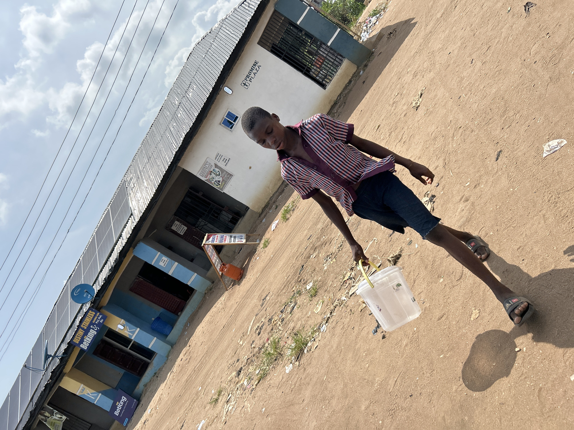 In Ihiagwa, Nigeria, a young boy, dressed in a striped short-sleeved shirt and dark shorts, carries a transparent plastic bucket with a yellow handle. He is walking on a dirt road. Behind him are buildings with corrugated metal roofs and signage, including "PROMISE PLAZA" and "DESTINY STAINLESS Betking & GAMING HUB Etc." There are litter and vegetation along the roadside. Electrical wires are visible against the sky.