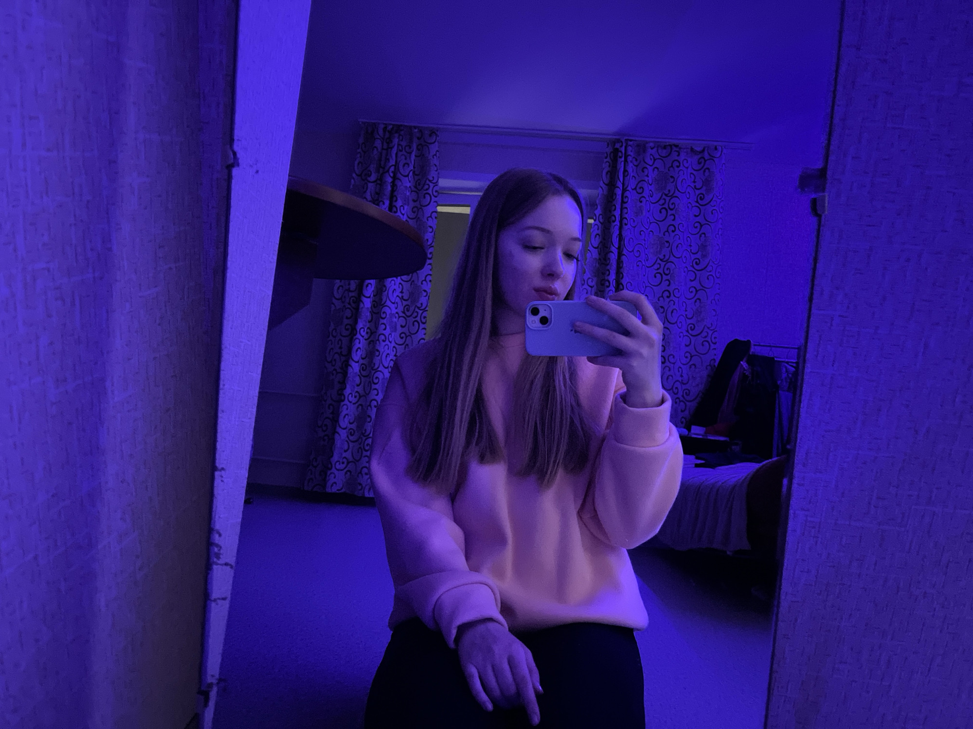 The photograph appears to be a selfie taken in a room illuminated by blue light, possibly from an LED source. The subject is a young woman, fair-skinned with long, light-brown hair, wearing a pink sweatshirt and dark-colored pants. She is holding a light-colored smartphone, presumably taking the picture.
The room has patterned curtains visible in the background, along with a partially visible bed and other furniture. The walls have a textured wallpaper. The photograph seems to have been taken in front of a mirror, as evidenced by the reflections on either side of the subject. Based on publicly available information, and assuming the photographer made no errors and no other data is available, it is not possible to confirm the location of this photo as Chaikovsky, Russia. The available image data does not explicitly contain information regarding the location.