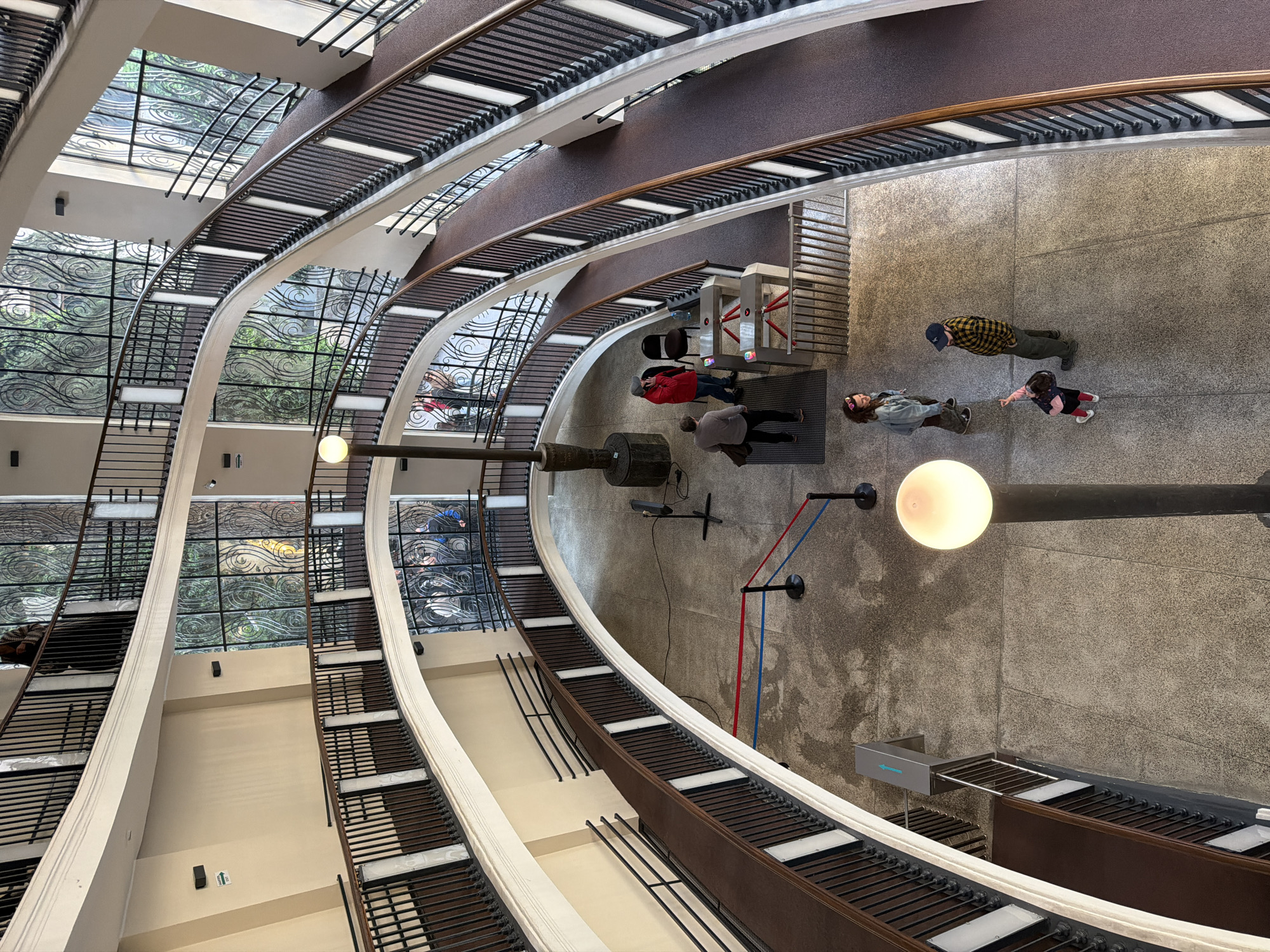 A high-angle shot shows the interior of a multi-story building in Tbilisi, Georgia, specifically the Bank of Georgia Headquarters, designed by architects George Chakhava and Zurab Jalaghania. The architecture features rounded balconies with dark brown metal railings and white structural supports that create a circular pattern around a central open space.
On the ground floor, several people are present. One group of people is near what appears to be a security checkpoint with metal turnstiles. Another group includes a man in a plaid shirt and a woman standing near a child. A lamp is visible on the wall.
The interior is well-lit, combining natural light coming through the glass windows with interior lighting. The ground floor is grey.