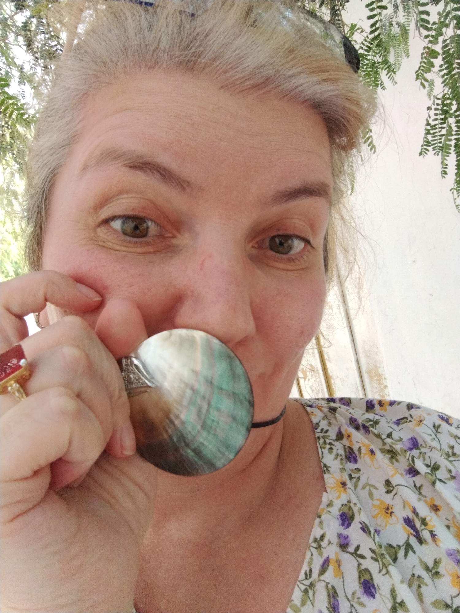 A selfie of a light-skinned woman with graying blonde hair. Her eyes are brown. She is wearing a floral-patterned white blouse with yellow and purple flowers. The woman is holding a large, iridescent seashell-like pendant up to her face. She is wearing two rings on her left hand: one with a large red stone and another with smaller stones. The background shows some greenery and a white wall or surface. The location is not determinable as Al Mas'oudi, United Arab Emirates.