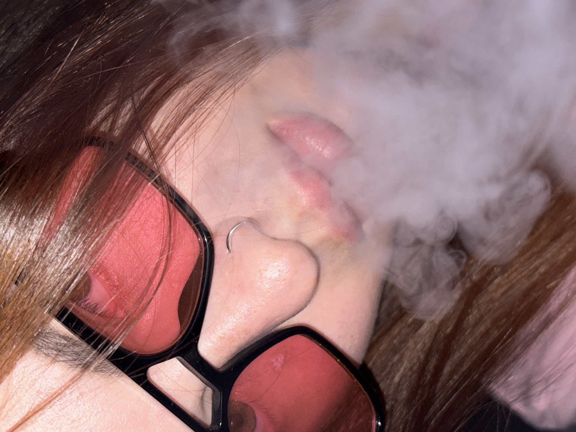 A close-up image shows a person's face partially obscured by a cloud of smoke. The person has light skin, brown hair, and is wearing sunglasses with dark frames and red lenses. A silver nose ring is visible. The person's lips are parted as if exhaling smoke. The background is out of focus, with some hair visible. The location is not discernible from the image. The city of Krasnodar, Russia, cannot be determined from this image.