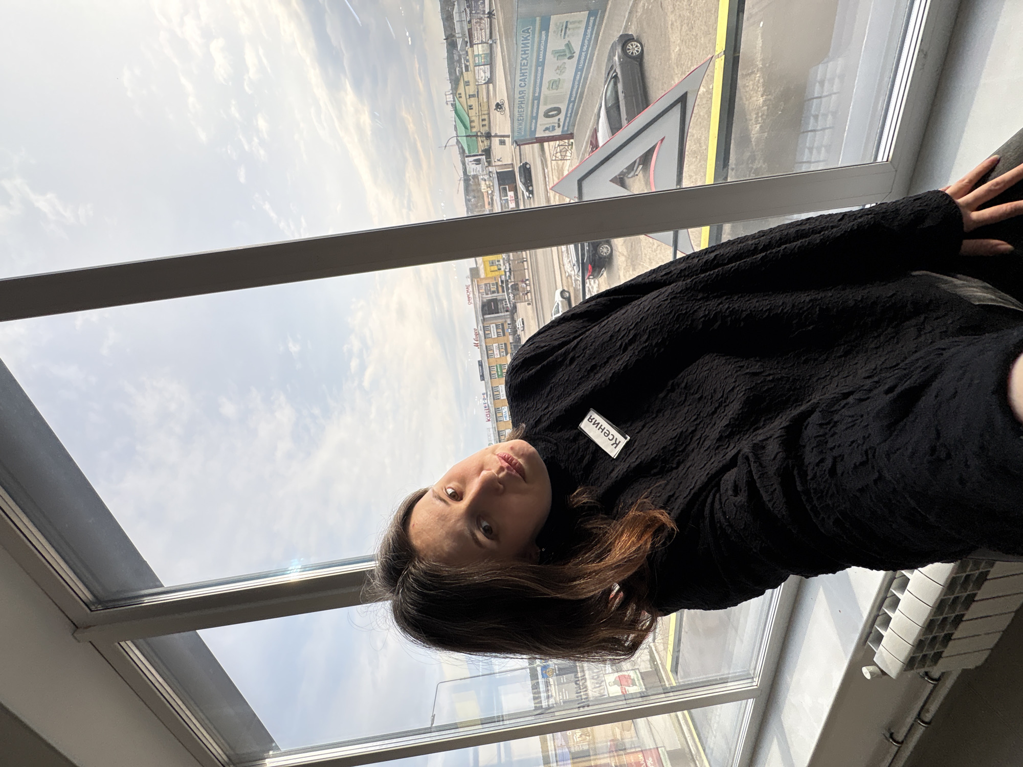 The image shows a woman wearing a black sweater with a name tag that reads "Ксения" (Ksenia) in Russian. She is positioned indoors near a large window overlooking a cityscape. The window provides a view of buildings, streets, vehicles, and signs. One visible sign indicates "ИНЖЕНЕРНАЯ САНТЕХНИКА" (Engineering Plumbing) in Russian, and the text "ОФИСМАГ" can also be seen. The environment suggests a location in the city of Chaikovsky, Russia. The woman is looking directly at the camera.