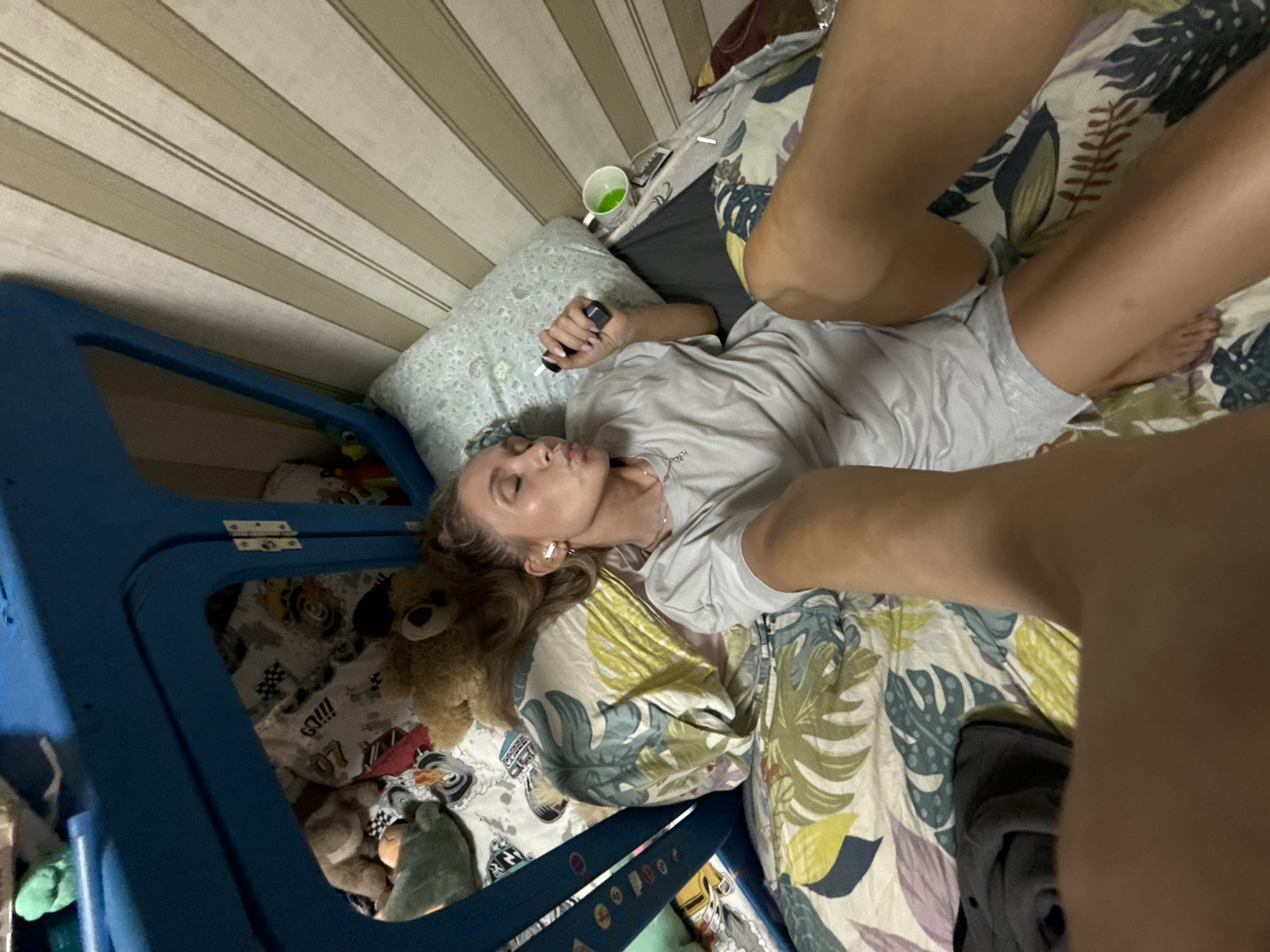 In Chaikovsky, Russia, a young woman with blonde hair, wearing a light gray t-shirt and shorts, lies on a bed with a colorful floral-patterned comforter. She is holding a black object in her right hand and has white earbuds in her ears. Her eyes are closed, and her lips are pursed. The room has striped wallpaper, and a blue play structure or bunk bed frame is visible to the left. Several stuffed animals are scattered on the bed, including a brown teddy bear. A green beverage is in a white mug on a surface next to the bed.