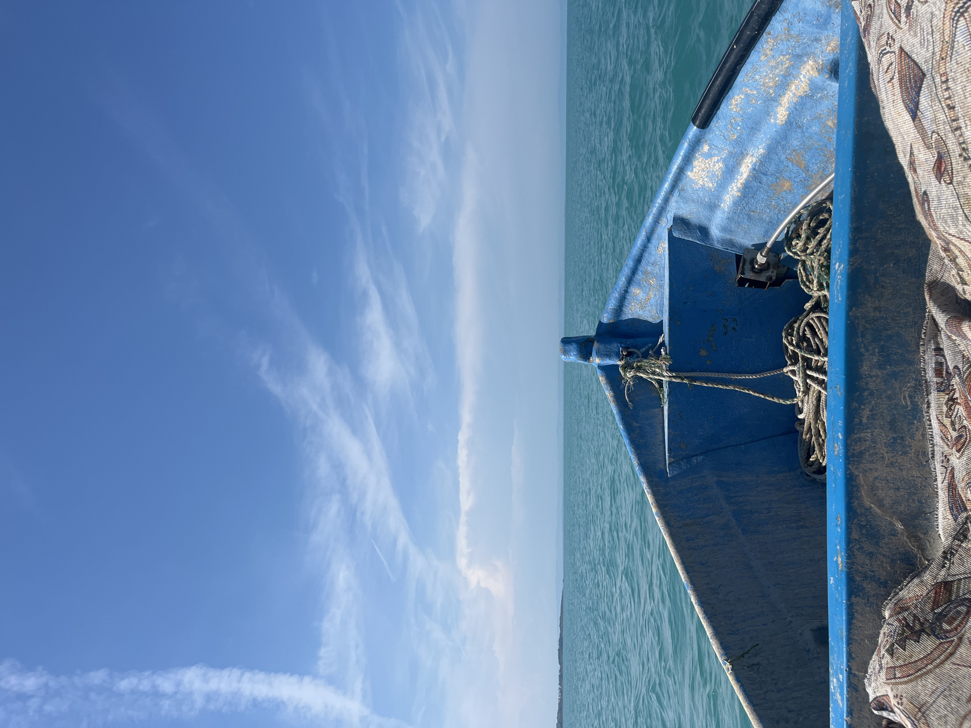 The image presents a first-person perspective from the bow of a small, weathered blue boat traversing open water. The boat's interior features coiled ropes near the bow and a patterned fabric, possibly a rug or blanket, covering the immediate foreground. The blue paint of the boat is chipped and faded in multiple areas, revealing lighter undertones and dirt accumulation.
The water, appearing in shades of turquoise and teal, exhibits minor ripples across its surface, extending to a distant horizon line. Along the left side of the horizon, a low-lying strip of land or an island is faintly visible. Above the horizon, the sky is a clear, light blue, populated by scattered white clouds. A distinct, elongated white cloud or contrail stretches vertically down the left side of the upper sky. Another, more voluminous white cloud mass is situated lower near the horizon, slightly to the left of the center. The lighting indicates bright daylight conditions.