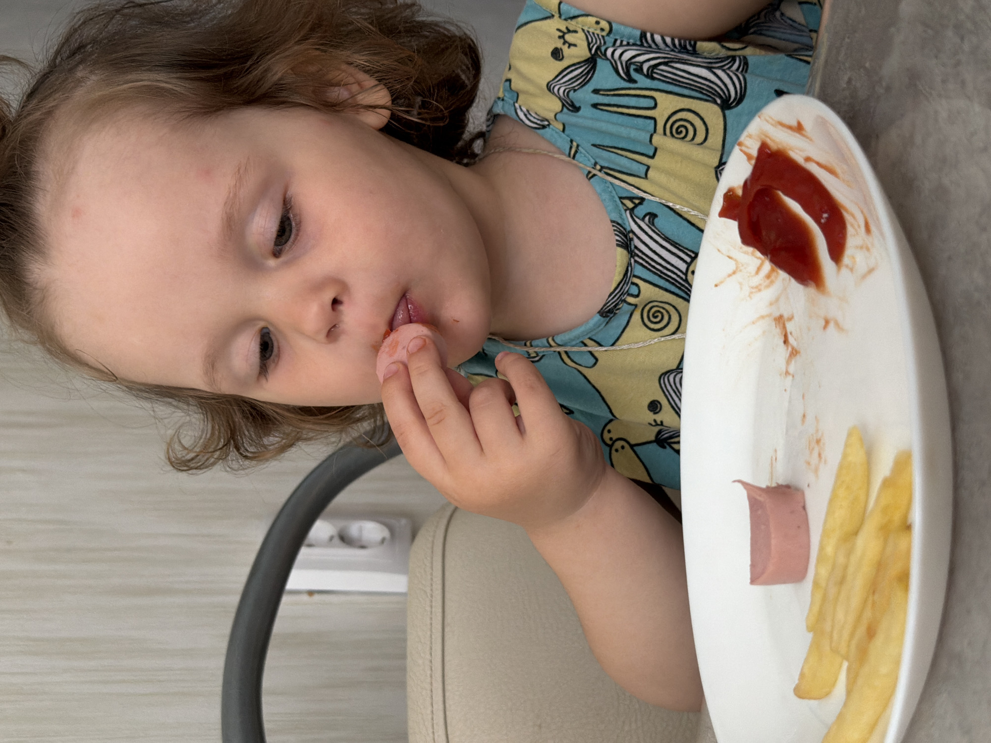 A young child with light skin and brown hair is sitting at a table with a white plate containing ketchup, fries, and a piece of sausage. The child is picking up a piece of food with their fingers and placing it in their mouth. The child is wearing a light blue and yellow shirt with animal designs. The child is seated in front of a gray wall with an electrical outlet. The scene appears to be indoors. The location information requested cannot be confirmed by visual analysis of the image.