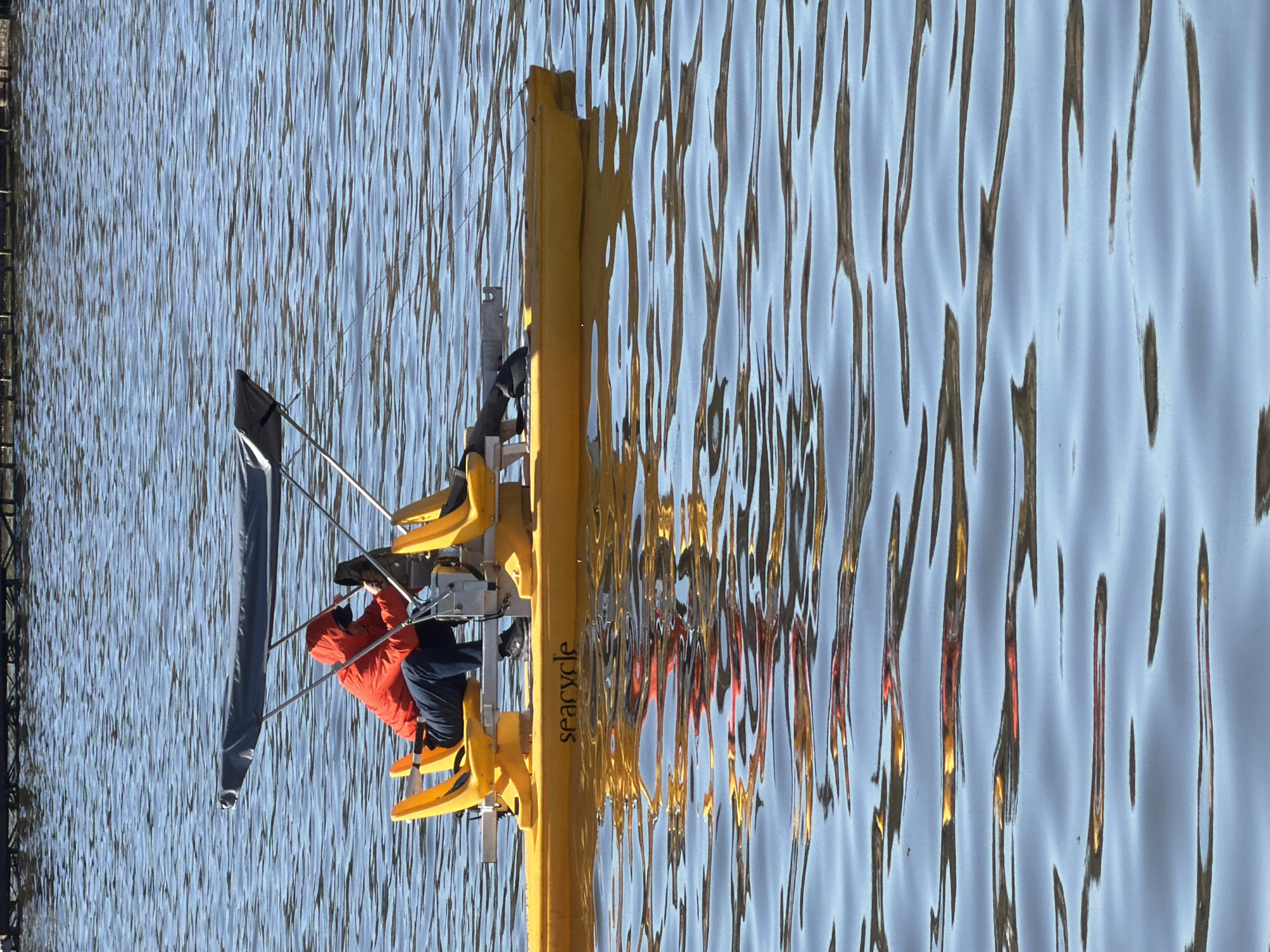 A wide shot captures a yellow "seacycle" watercraft on the water in Votkinsk, Russia. The seacycle is equipped with yellow seats and a gray sail. A person in an orange hooded jacket and another person in dark clothing are seated on the cycle. The surface of the water is rippled and reflects the seacycle and sky. A dark fence is visible on the left edge of the frame.