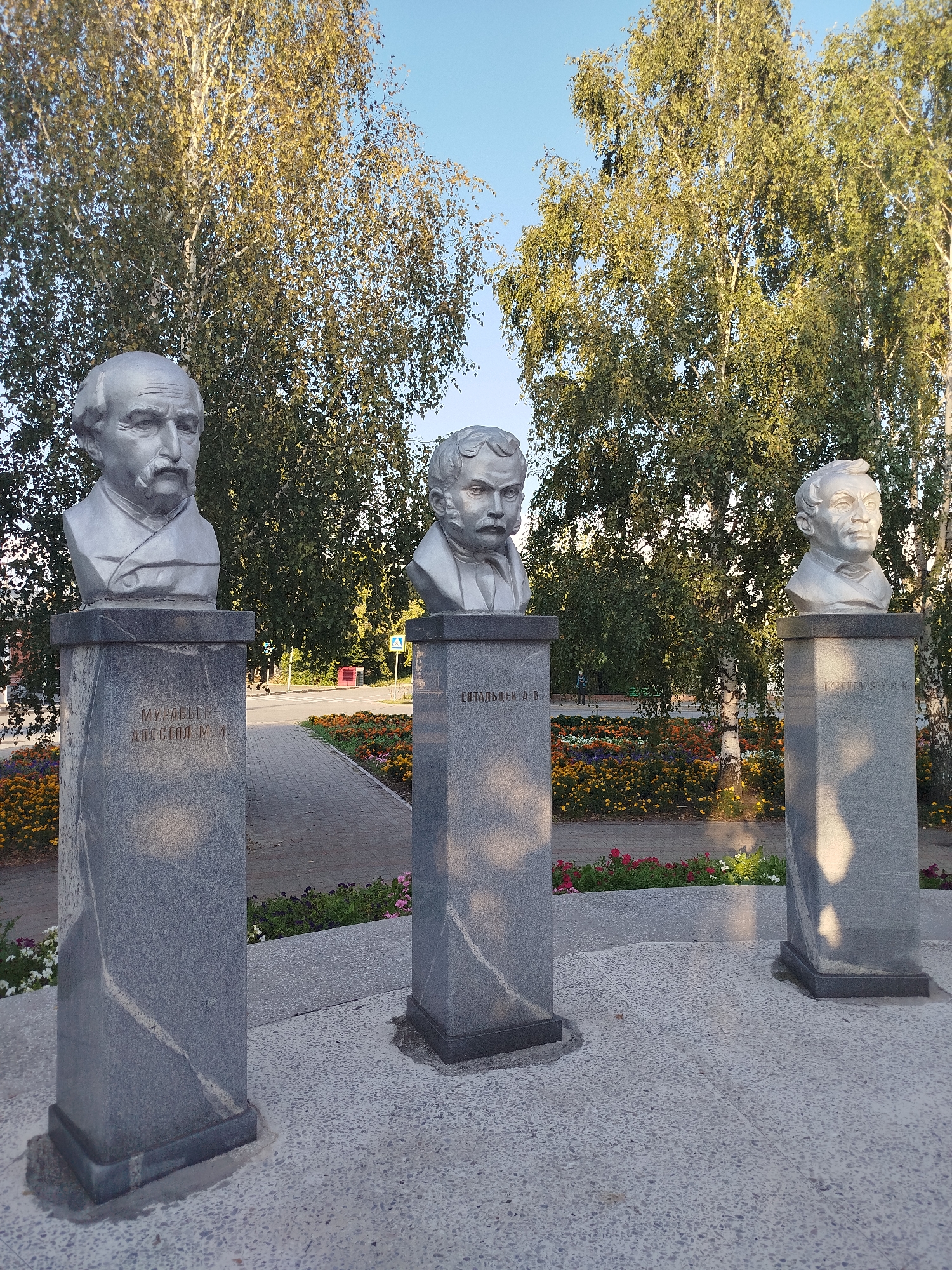Three granite busts of historical figures stand on granite pedestals in a park setting. The busts depict men with distinct facial features and hairstyles. The pedestals are inscribed with text. In the background, there are tall trees with green and yellow leaves, a paved walkway, and flowerbeds with colorful flowers. The sky is clear and blue. This scene is located in the city of Yalutorovsk, Russia, at the memorial to the Decembrists, specifically honoring Mikhail Muravyov-Apostol, Ivan Pushchin, and Alexander Odoevsky.
