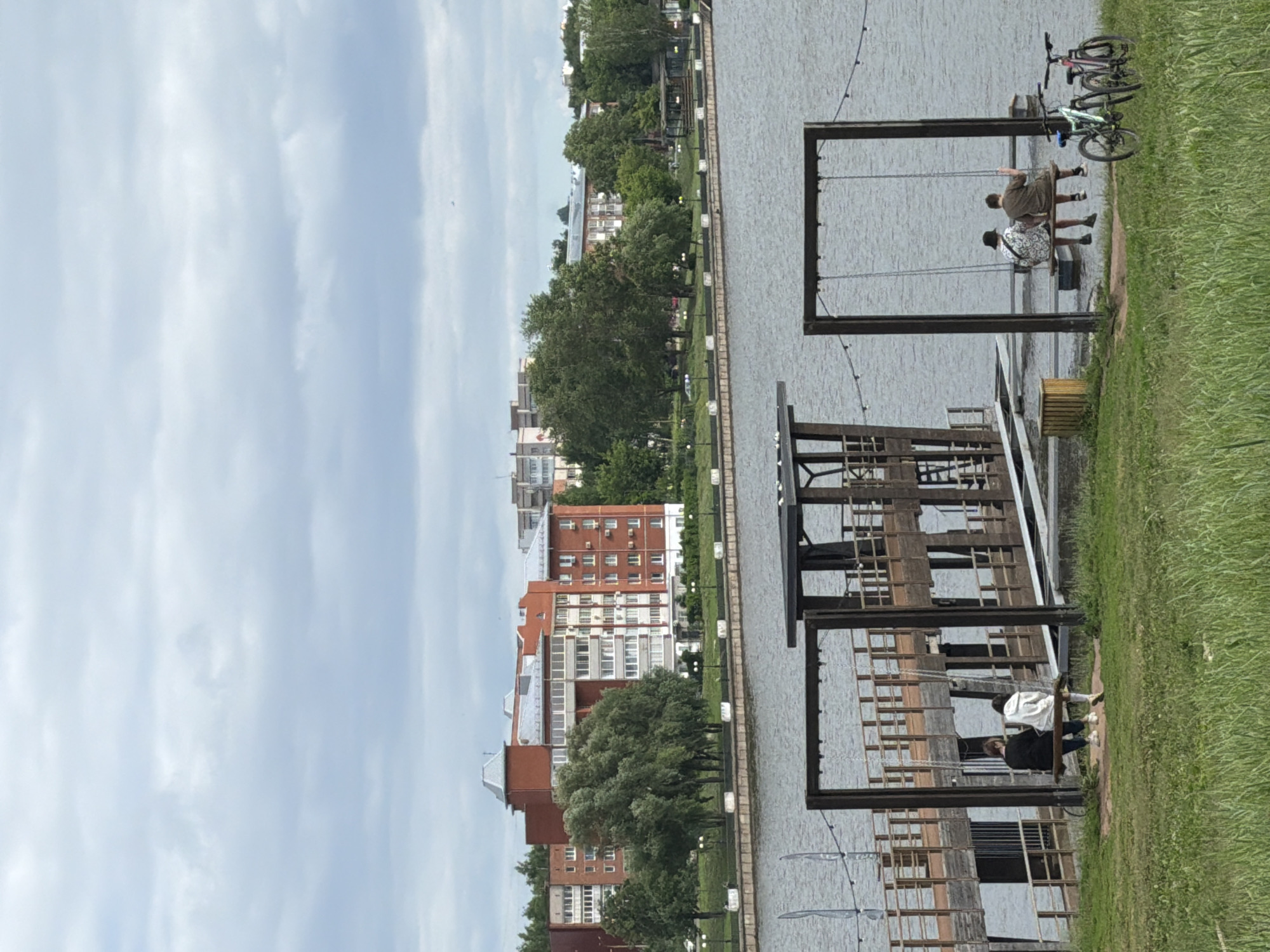A wide shot captures a waterside scene in Votkinsk, Russia. The foreground features lush green grass with two sets of swings. On the left swing, two individuals sit facing the water. On the right swing, two individuals are seated, also facing the water. Two bicycles, one green and one pink, are parked on the grass to the right of the right swing. In the midground, a wooden pier extends over the water. Further back, a body of water stretches towards a tree-lined embankment. Beyond the trees, several multi-story brick apartment buildings are visible, indicative of urban development. The sky above is overcast with a layer of gray clouds.