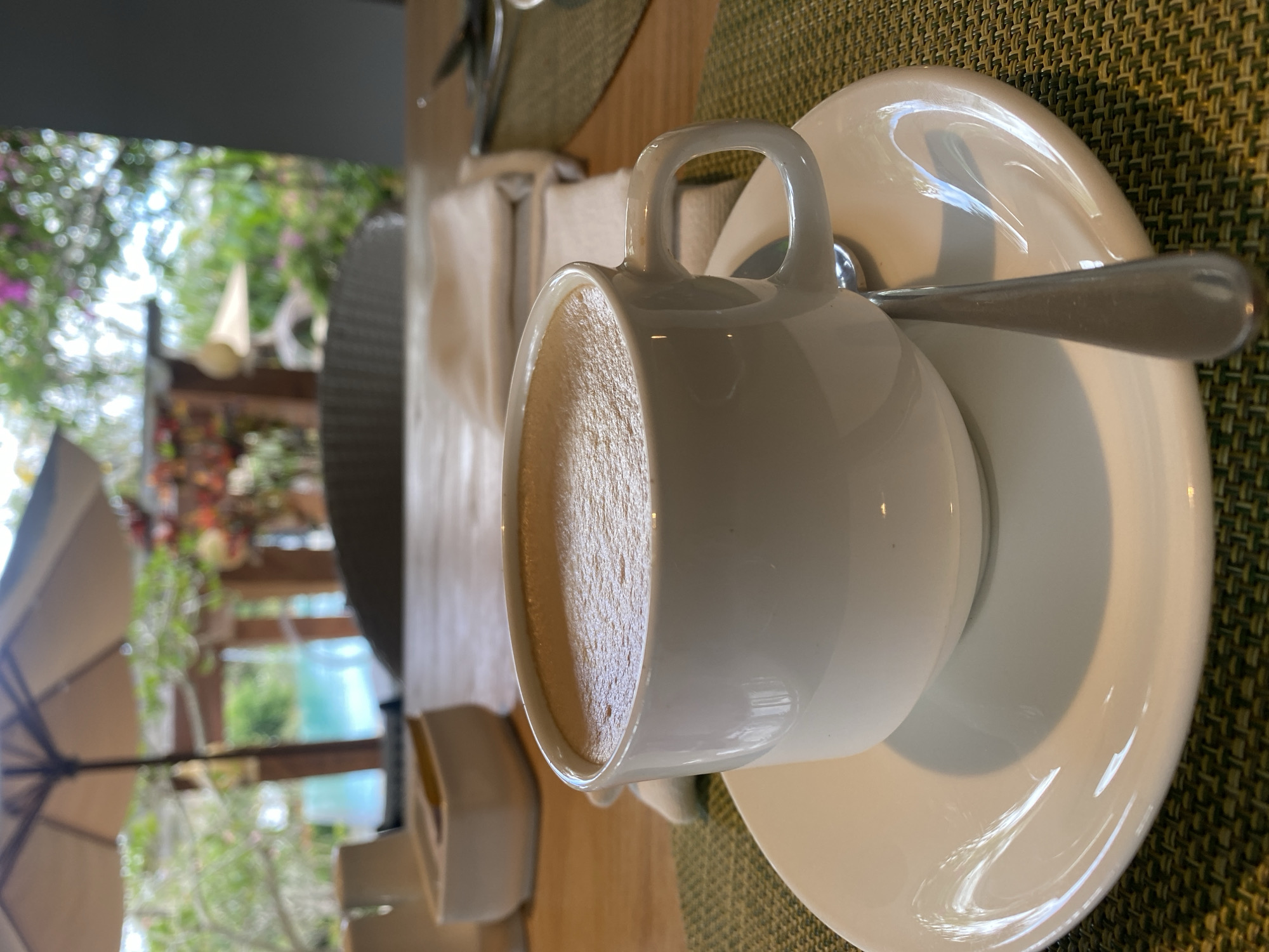 A close-up, low-angle shot shows a white cup of coffee with foam on a saucer, placed on a green woven placemat on a wooden table. A spoon rests on the saucer. In the blurred background, an outdoor seating area with umbrellas and foliage suggests a resort or restaurant in Kuta Selatan, Indonesia. A swimming pool is visible in the distance.