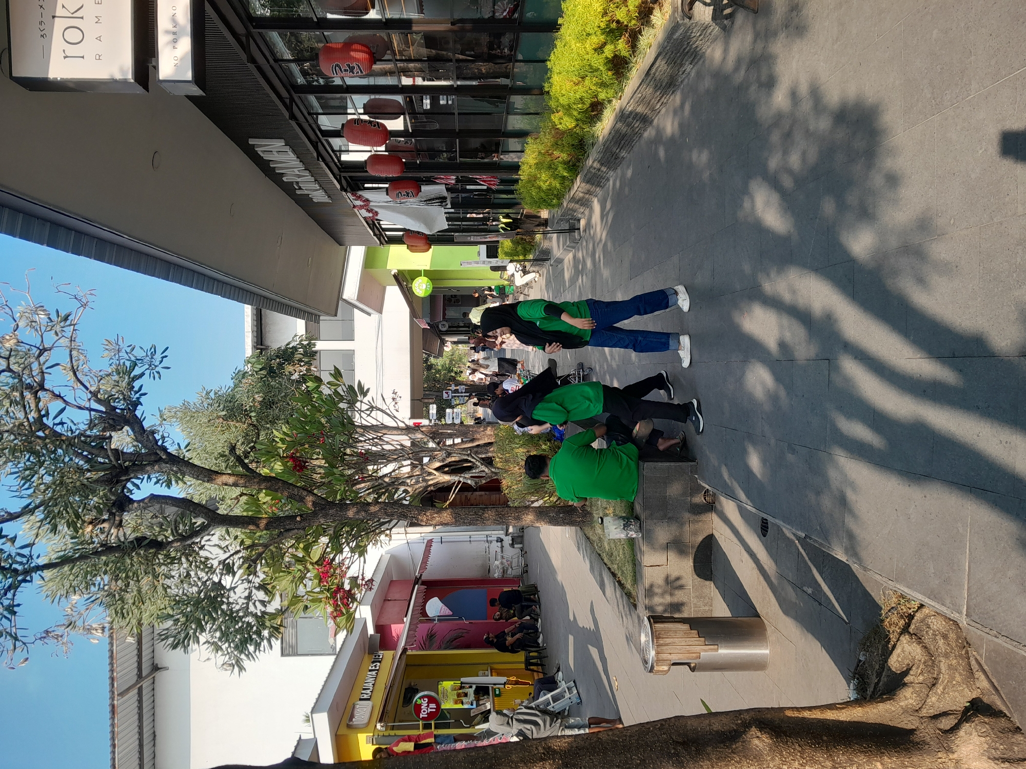 The image depicts an outdoor pathway in Sempusari, Indonesia, likely a commercial area or mall. The pathway is paved with grey square tiles and lined with greenery on the right. Several people are present. In the foreground, two individuals wearing green shirts and dark pants are standing and sitting on a grey stone bench. One person is looking towards the left, and the other is seated. To the left of the bench is a cylindrical trash bin with a wooden slatted exterior. Further down the pathway, a woman wearing a black hijab and a green shirt with blue jeans walks away from the camera. In the background, several shops line the pathway. On the left, a yellow storefront with a red and white awning has signage that reads "TONOT RAJAANYA ESTEH". Several people are seated outside this establishment. On the right, a building with large glass windows and a dark facade has Japanese lanterns hanging outside, with signage that includes "ROK... RAMEN". The sky is clear and blue, and shadows from trees are cast on the pathway, indicating a sunny day.