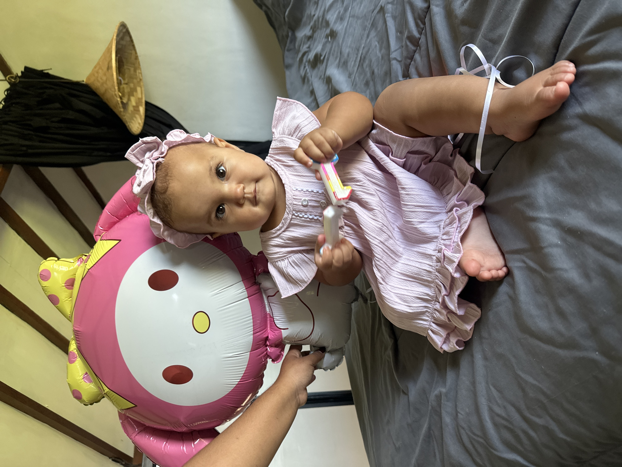 An infant girl wearing a pink ruffled dress and matching headband sits on a bed covered with a dark gray comforter. She is holding a colorful toy in her hands. To her left, a person's arm is visible, holding a large pink and yellow My Melody balloon. In the background, a black curtain hangs, and a woven conical hat hangs from a beam. The scene appears to be indoors, possibly in a bedroom.