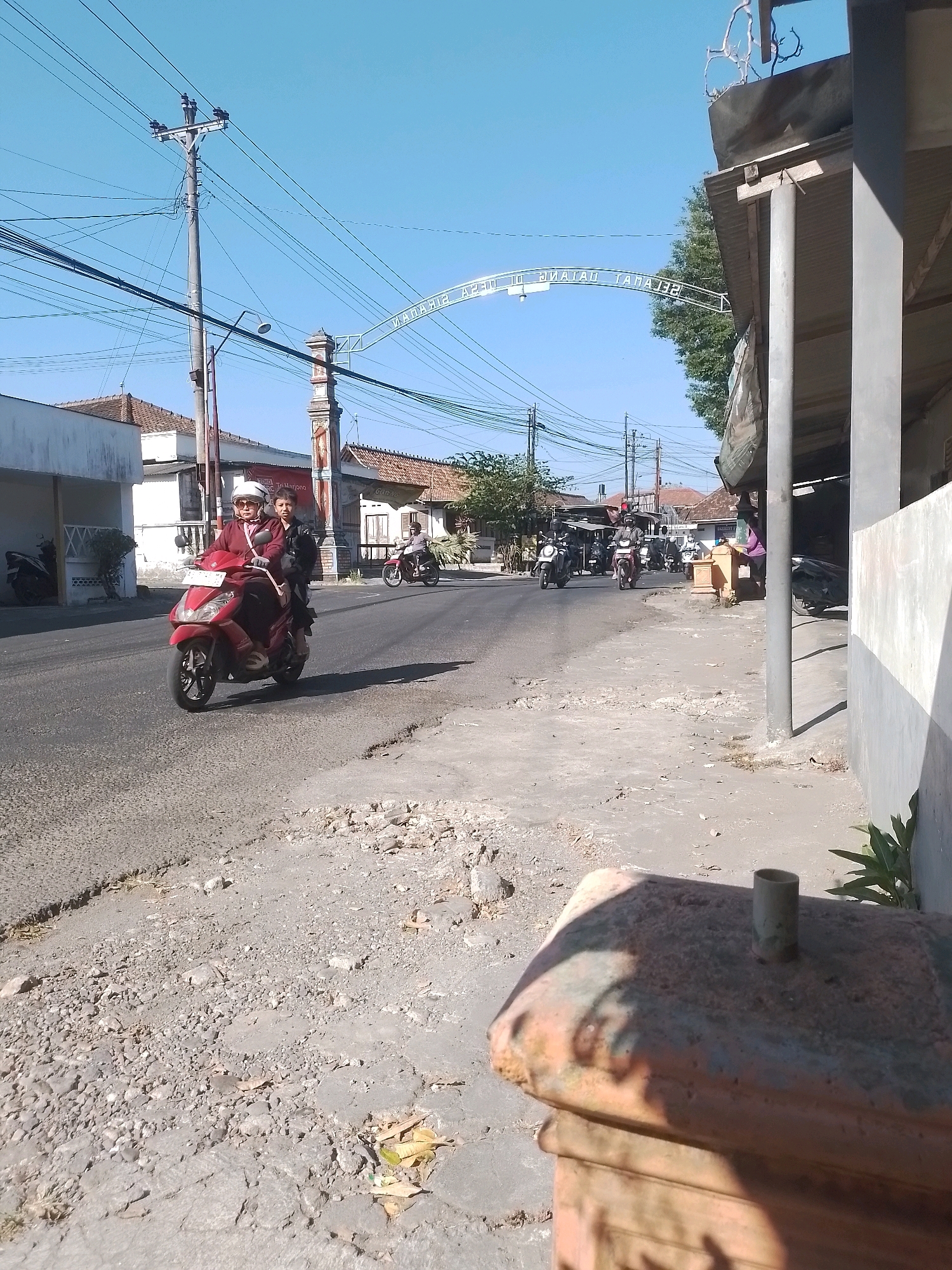 The image shows a street scene in Wonosari, Indonesia, under a clear blue sky. In the foreground, on the right side, is a partially visible rusty metal structure, possibly a post or a monument. The street itself is paved with asphalt and shows signs of wear and tear, with debris and gravel scattered on its surface.
On the left side of the street, there is a red motorcycle carrying two people, a woman in front wearing a helmet and a maroon jacket, and a passenger behind her wearing a black jacket. The motorcycle is in motion, heading towards the viewer. Further down the street, more motorcycles and a few people are visible. There are buildings lining the street, some appearing to be houses with tiled roofs and white walls.
In the background, an archway spans the road with some text inscribed on it. Several utility poles with numerous wires are also present. The overall scene depicts a typical day on a street in a residential or commercial area of Wonosari.