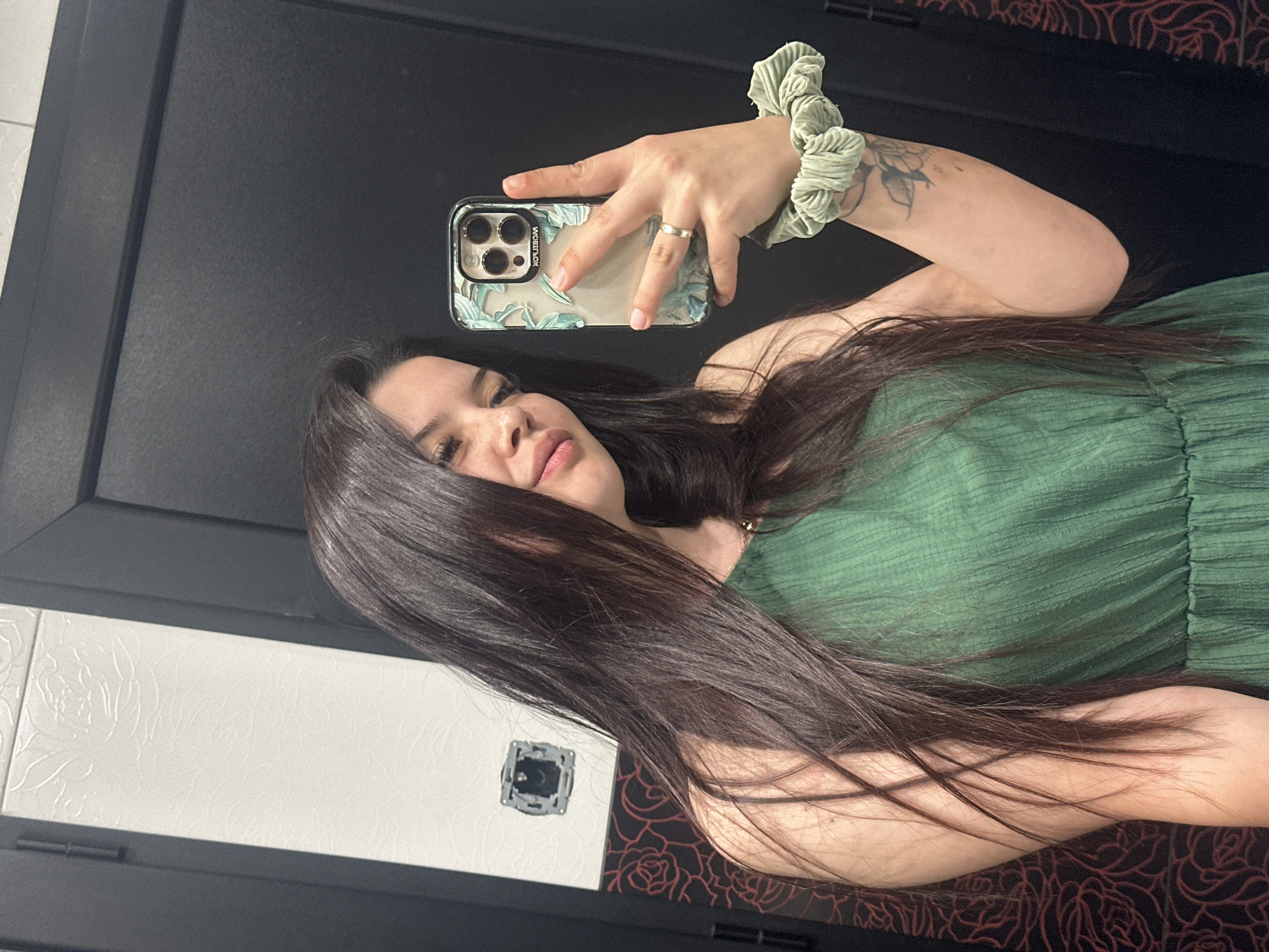 A young woman with long, dark hair is taking a selfie in a dimly lit room. She is wearing a green dress and has a phone in her right hand, which is decorated with a floral case. On her left wrist, she wears a light green scrunchie. A ring is visible on her right ring finger. The background features a white textured wall with a light switch, and a black door. There is also a section of wall with a dark background and a repeating red floral pattern. The scene appears to be an indoor setting, possibly a bathroom or dressing area, though no specific location in Sliven, Bulgaria, can be determined from the image alone.