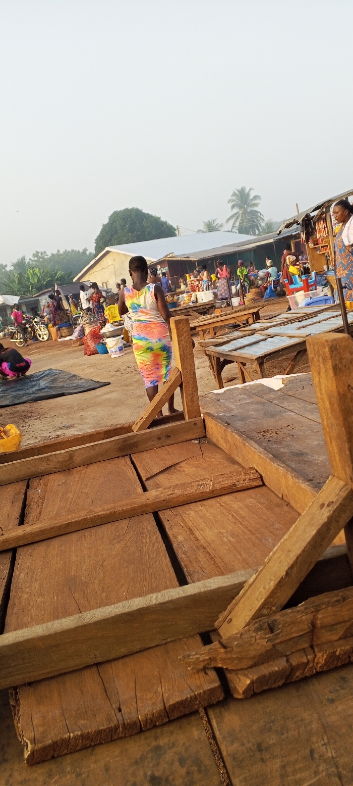 A vibrant marketplace scene unfolds under a hazy sky. Wooden tables dominate the foreground, hinting at stalls. A woman in a colorful dress walks away from the viewer. In the background, figures gather near buildings and other market stalls, some shaded by palm trees. A motorcycle is visible among the crowd. The scene suggests a bustling local market in a tropical setting.