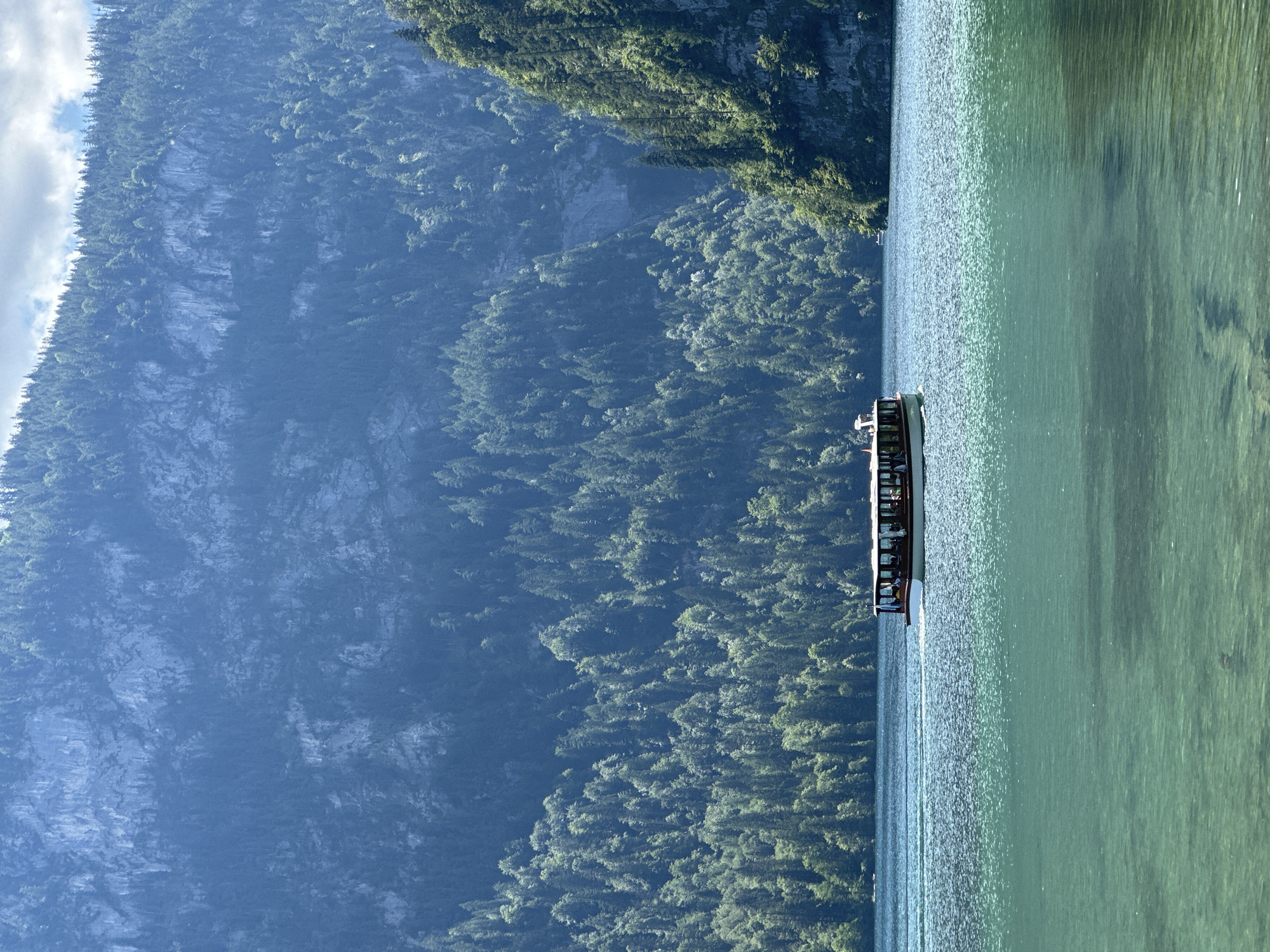 This image captures a serene view of Königssee lake in Schönau am Königssee, Germany. A white and brown passenger boat with several people visible inside is navigating the crystal-clear, turquoise waters. The boat is moving from left to right, leaving a gentle wake. The lake is surrounded by steep, densely forested mountains that rise dramatically in the background. The trees cover the mountain slopes from the water's edge to the rocky peaks. The sky above is partly cloudy. The overall scene conveys a sense of tranquility and natural beauty.