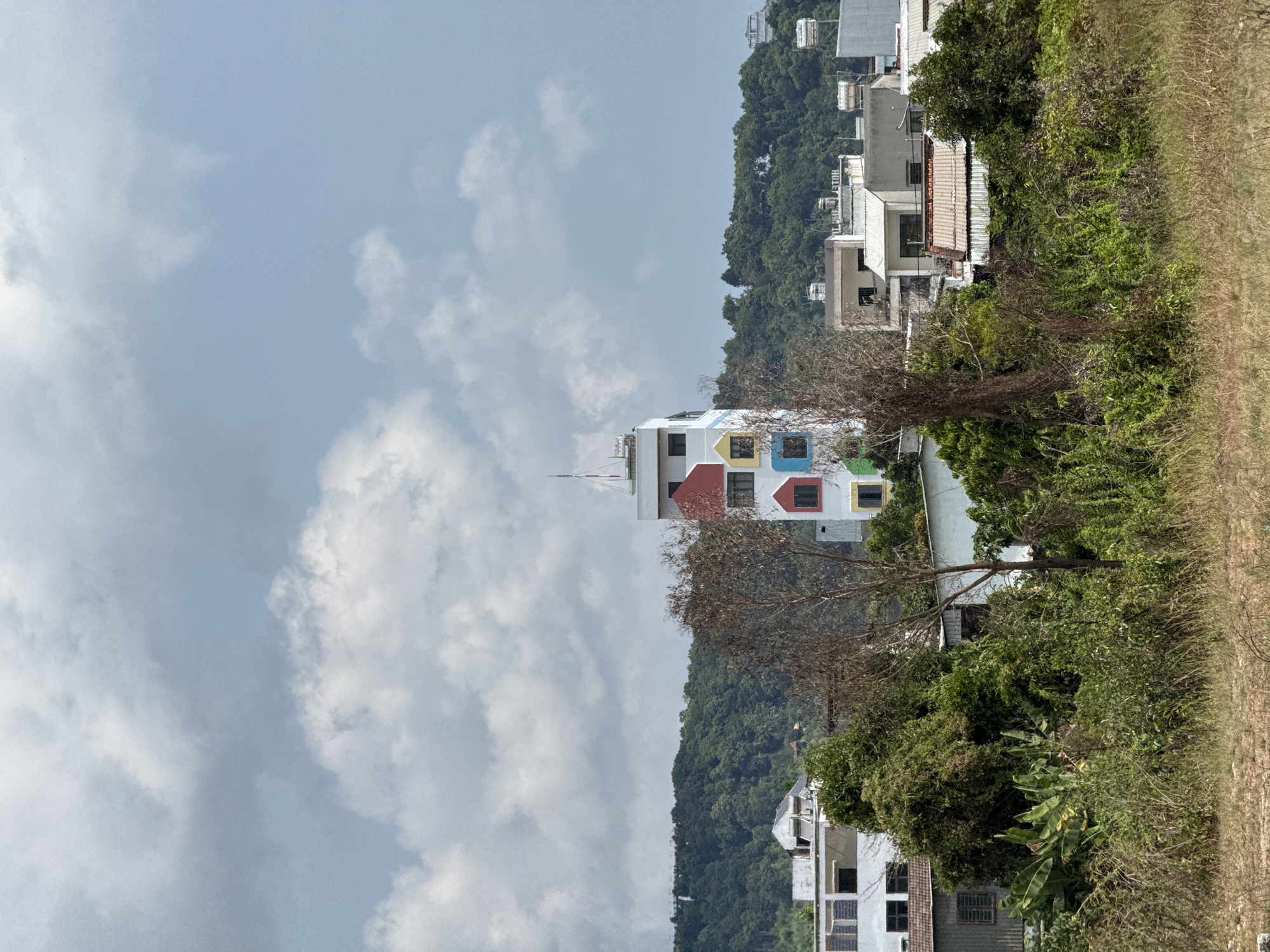 The image shows a tall, white building with colorful window frames (red, yellow, blue, green) in Phú Quốc, Vietnam. The building stands out against a backdrop of green hills and a cloudy sky. In the foreground, there are trees and vegetation, and in the background, other buildings are visible on the hillside.