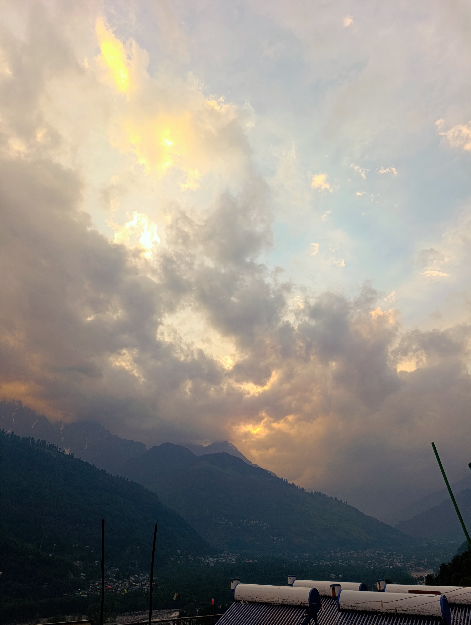 The image depicts a serene sunset over the town of Vashisht, India, nestled in the Himalayas. The sky is a canvas of dramatic clouds, illuminated by the warm glow of the setting sun, which peeks through the cloudy expanse, casting golden hues across the sky. The mountainous terrain of the Himalayas forms a majestic backdrop, with the peaks shrouded in a soft, dusky light. Below the mountains, the town of Vashisht is visible, with its buildings and lush greenery creating a stark contrast against the rugged landscape. In the foreground, there are solar water heaters installed on a rooftop, suggesting the presence of residential or commercial buildings. The overall scene evokes a sense of tranquility and natural beauty, characteristic of the Himalayan region.