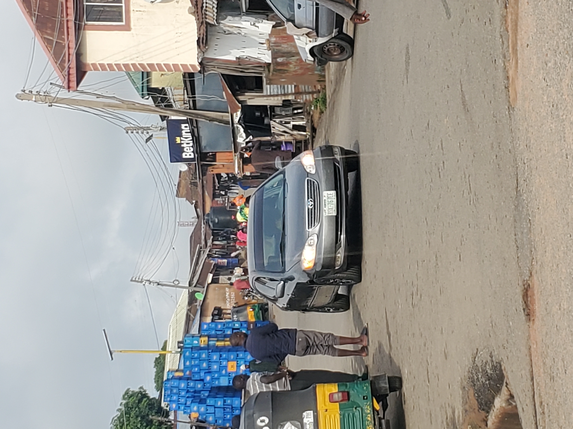 The image depicts a street scene in Jos, Nigeria. The perspective is from a slightly elevated angle, looking down onto a paved road lined with various shops and structures. The scene appears to be a bustling marketplace or commercial area.
In the foreground, a dark-colored sedan is parked or moving slowly on the right side of the road. Two men are visible near the car. One man, wearing a dark blue jacket and grey shorts, is standing and appears to be looking towards something out of frame. Another man, wearing a striped shirt and dark pants, is partially obscured by the car, standing next to a yellow and green tricycle, commonly known as an "Okada" or "Keke" in Nigeria, which is a form of public transportation. A stack of blue crates, possibly containing beverages, is prominent on the left side of the image, suggesting a commercial activity.
Behind the car and the men, the street is lined with buildings and makeshift stalls. Some of these structures appear to be made of corrugated metal and wood, indicating a developing or informal commercial setting. A sign with the text "BetKing" is visible on a pole, suggesting a betting shop or service. Overhead, a complex network of electrical wires crisscrosses the sky, a common sight in many urban areas in Nigeria. The sky is overcast, suggesting a cloudy or possibly impending rainy weather.
The overall atmosphere appears to be one of everyday commerce and street life. The time of day is indeterminate, but the lighting suggests it could be daytime. The presence of the tricycle and the general streetscape are strong cues to the location being in Nigeria.