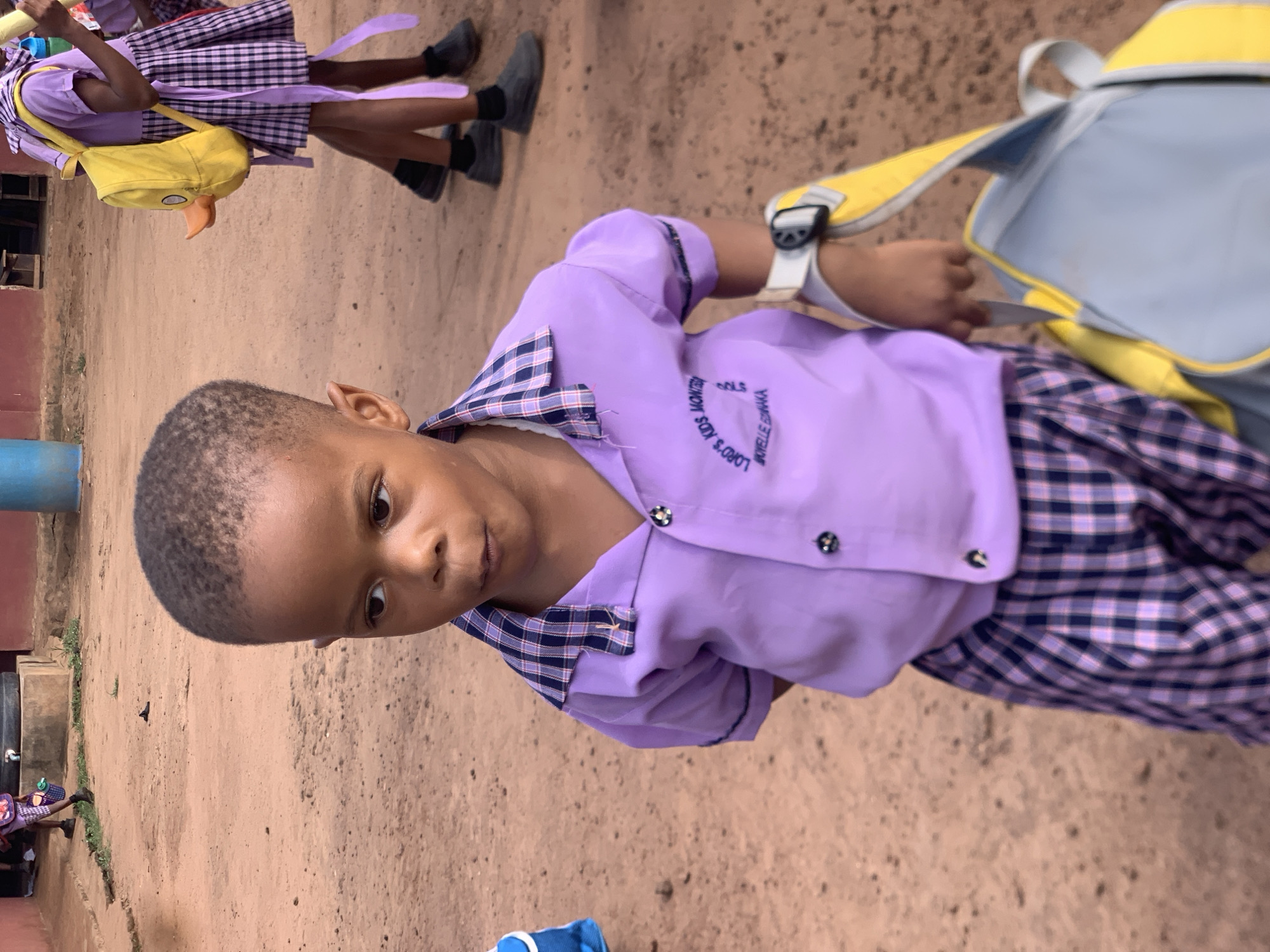 The image shows a young boy in the foreground wearing a lavender school uniform shirt and plaid shorts. The shirt has a dark blue and white checkered collar and trim on the sleeves, with two black buttons on the front. A white watch with a black strap is visible on his left wrist. He is holding a gray and yellow duffel bag with a yellow strap. In the background, another child in a similar uniform is visible carrying a yellow backpack shaped like a duck. The ground is dirt, suggesting an outdoor setting, possibly a schoolyard or a rural area. The overall scene appears to be in Ukwu-Abwa, Nigeria, based on the provided information.