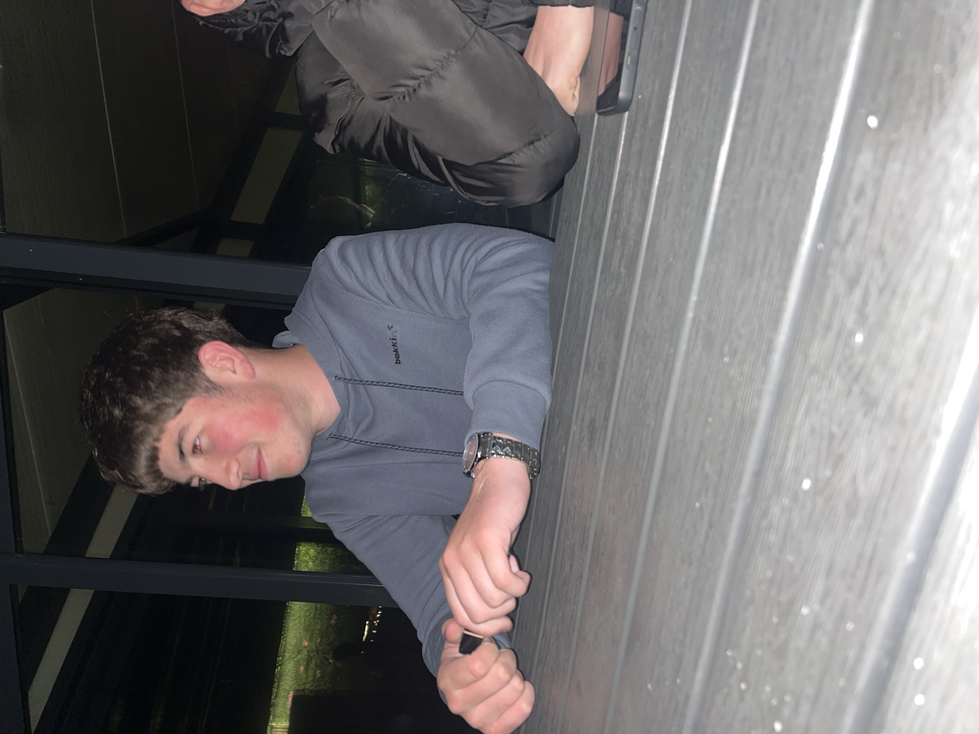 The image, originally oriented 90 degrees counter-clockwise, depicts two individuals in what appears to be an evening or nighttime setting in Kochan, Bulgaria.
In the foreground, a young man is visible from the chest up, positioned on the left side of the frame. He has short, dark brown hair and noticeably reddish cheeks. He wears a grey hooded sweatshirt with the text "bekkin" visible on the upper left chest area. On his left wrist, he sports a metallic watch with a dark face. His hands are clasped together, holding a small, dark cylindrical object, possibly a lighter or a vape device, and he is looking down at it with a focused expression.
To the right and slightly above the foreground man, the lower body and legs of another person are partially visible. This individual is wearing black, possibly puffy or padded, trousers. A black smartphone case is visible near their leg, suggesting the phone is resting beside them.
The setting includes a grey, ribbed surface in the right half of the image, resembling composite decking or an outdoor wall. Small, bright specks are visible on this surface, which could be water droplets or reflections. Behind the foreground man, there is a dark, reflective panel, likely a large window or glass partition, showing distorted reflections of green foliage and distant lights, indicating it is dark outside. The overall ambiance suggests a casual, nocturnal gathering.