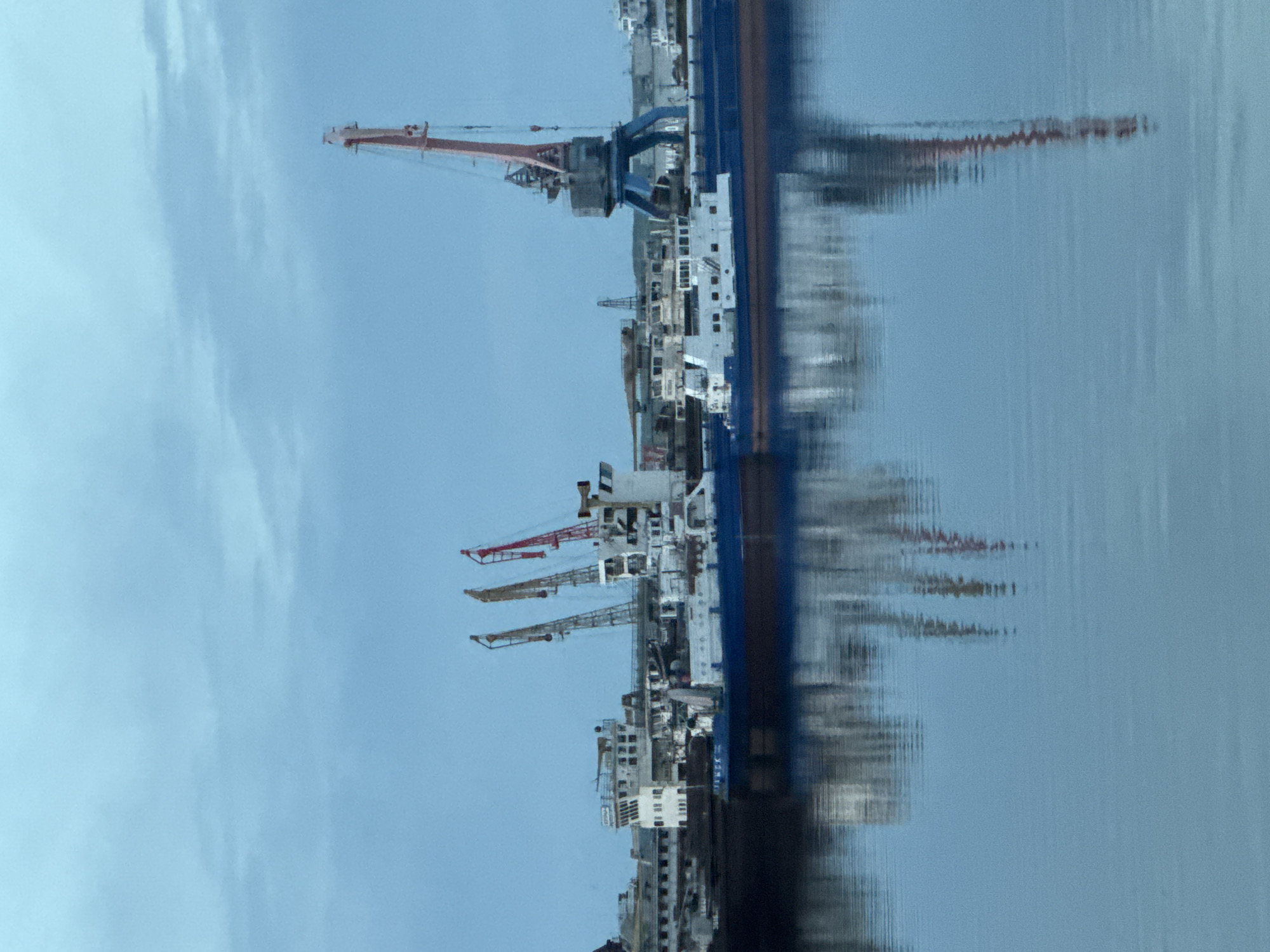 The image depicts a large vessel docked at a port in the city of Chaikovsky, Russia. The vessel, likely a cargo or transport ship, is lined with several cranes, suggesting industrial or commercial activity. The scene includes reflections on the water, indicating a calm or gently moving water surface. The sky is partially visible, appearing overcast or lightly clouded. There are no people visible in the image.