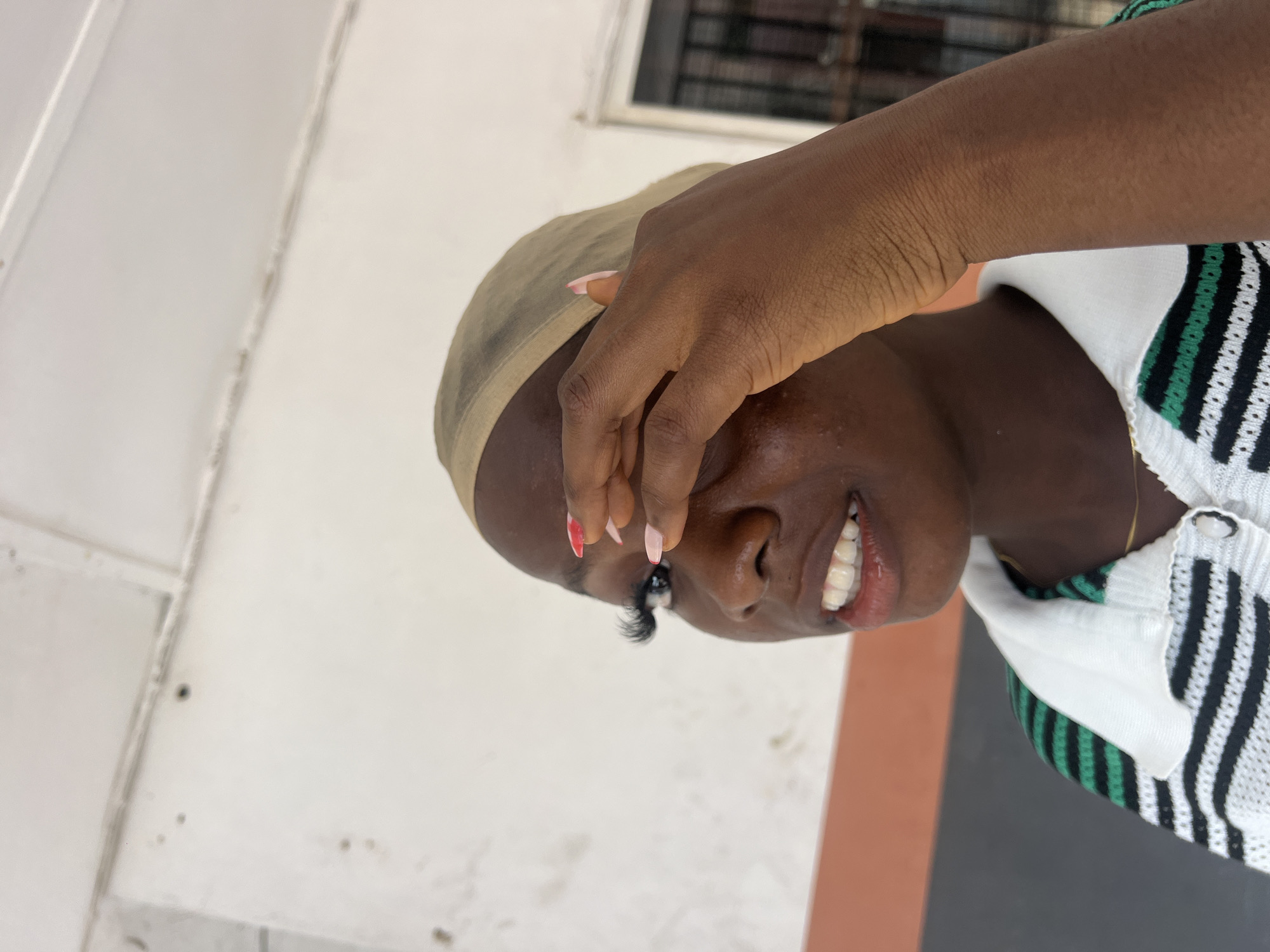 A close-up, eye-level, medium shot captures a young Black woman's face and upper body in what appears to be an outdoor urban setting, possibly Nkpor, Nigeria. She is wearing a tan wig cap that covers her hair. Her left eye is partially obscured by her right hand, which has long, bright pink acrylic nails with white tips. She has dramatic false eyelashes and her mouth is slightly open, revealing white teeth. She is wearing a collared, striped sweater with alternating bands of black, white, and green. The background shows a textured, off-white wall with horizontal lines and a dark horizontal band below. A portion of a barred window or gate is visible to the right. The lighting suggests it is daytime.