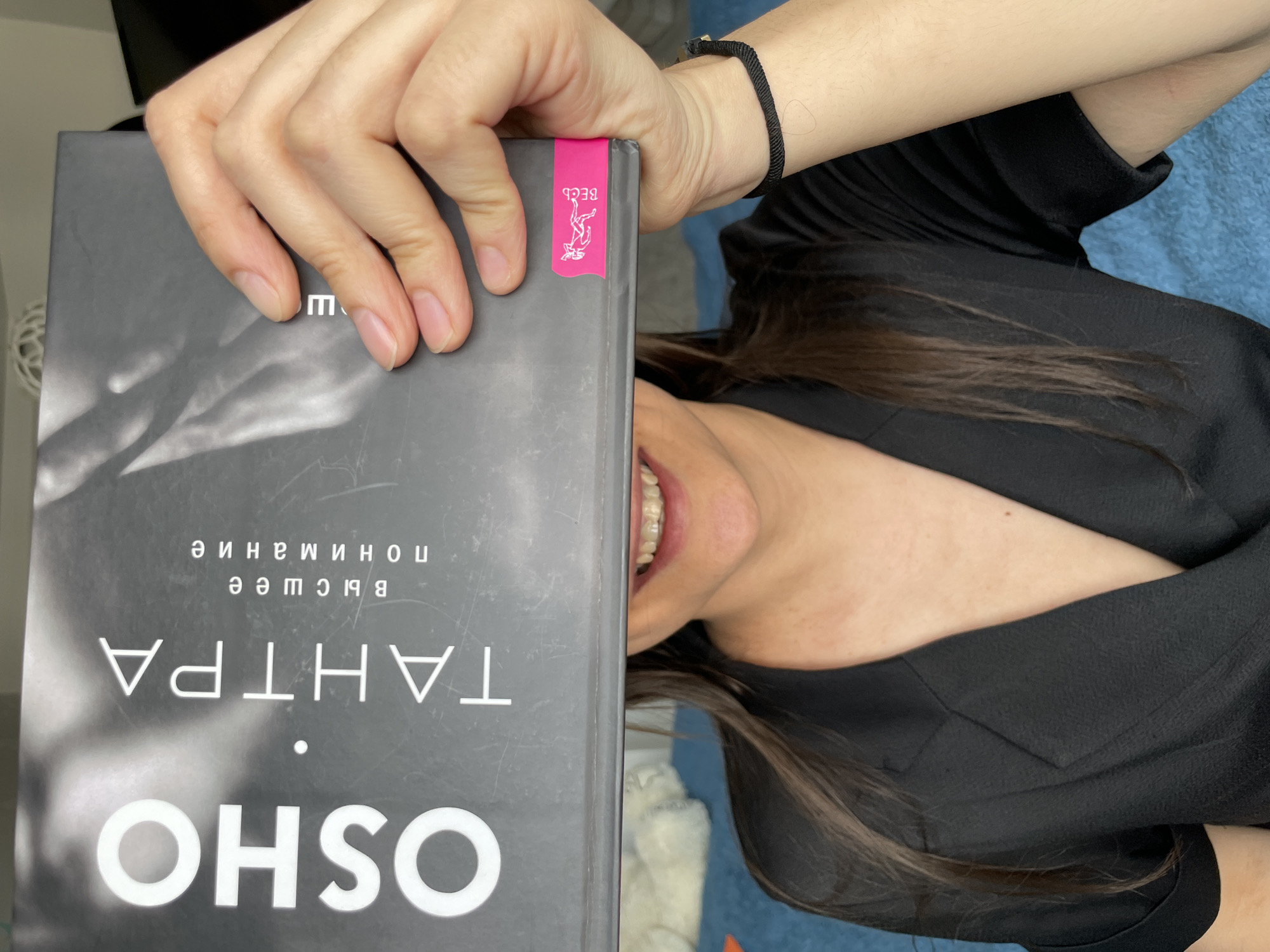 A person is holding a book titled "OSHO TANTRA" in front of their face, partially obscuring it. The book has a gray cover with white text in both English and a Cyrillic script. The person has fair skin, brown hair, and is wearing a black top. A black bracelet is visible on the person's wrist. The background appears to be a blue, textured surface.