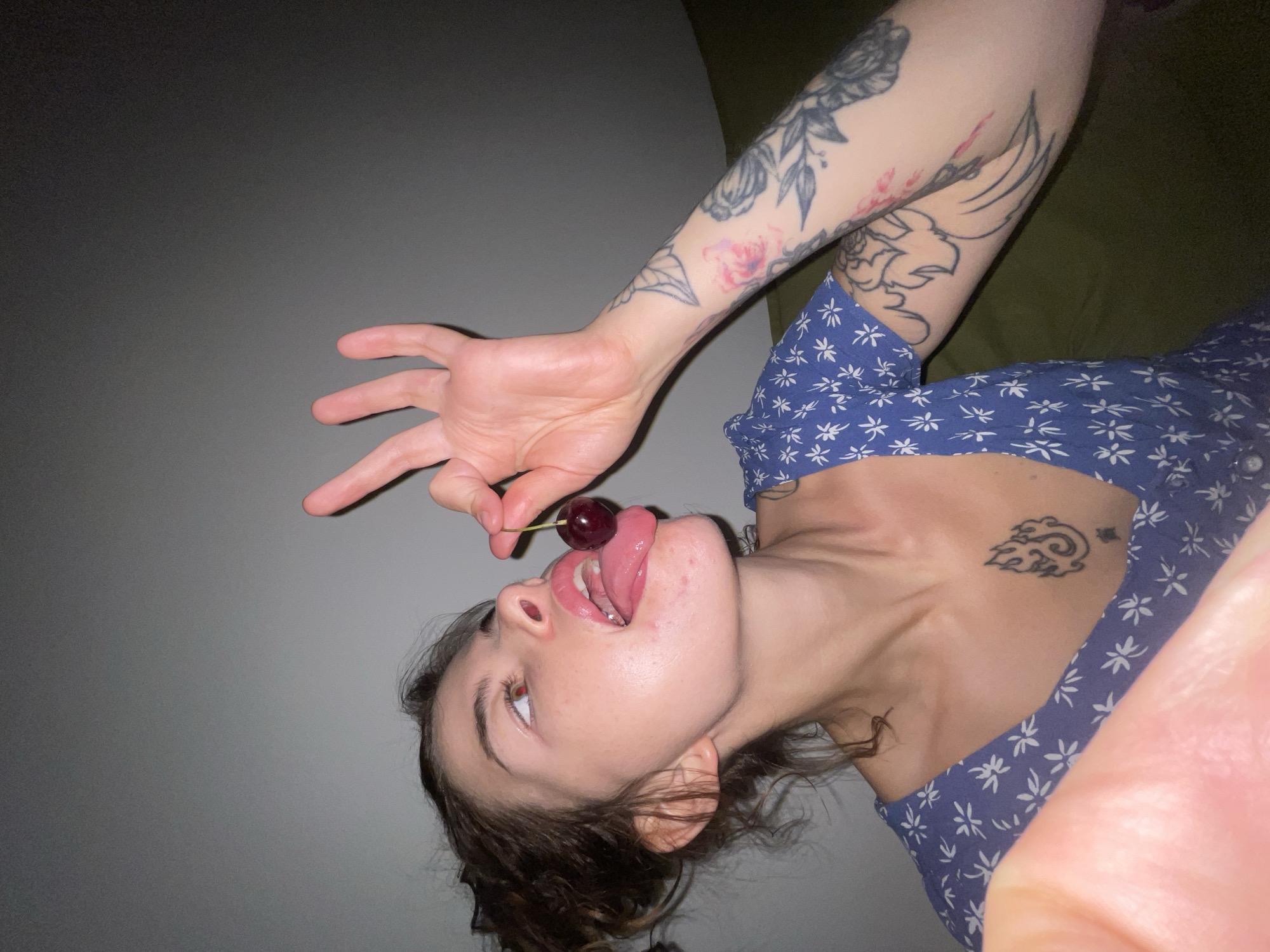 A young woman with dark, curly hair is holding a cherry with the stem between her fingers, bringing it towards her open mouth, with her tongue sticking out. She is wearing a blue dress with a white floral pattern. She has tattoos on her left arm and chest. The background is a plain, grey wall. The lighting is dim and appears to be artificial. The image does not contain any information that would indicate it is located in Boreti, Montenegro.