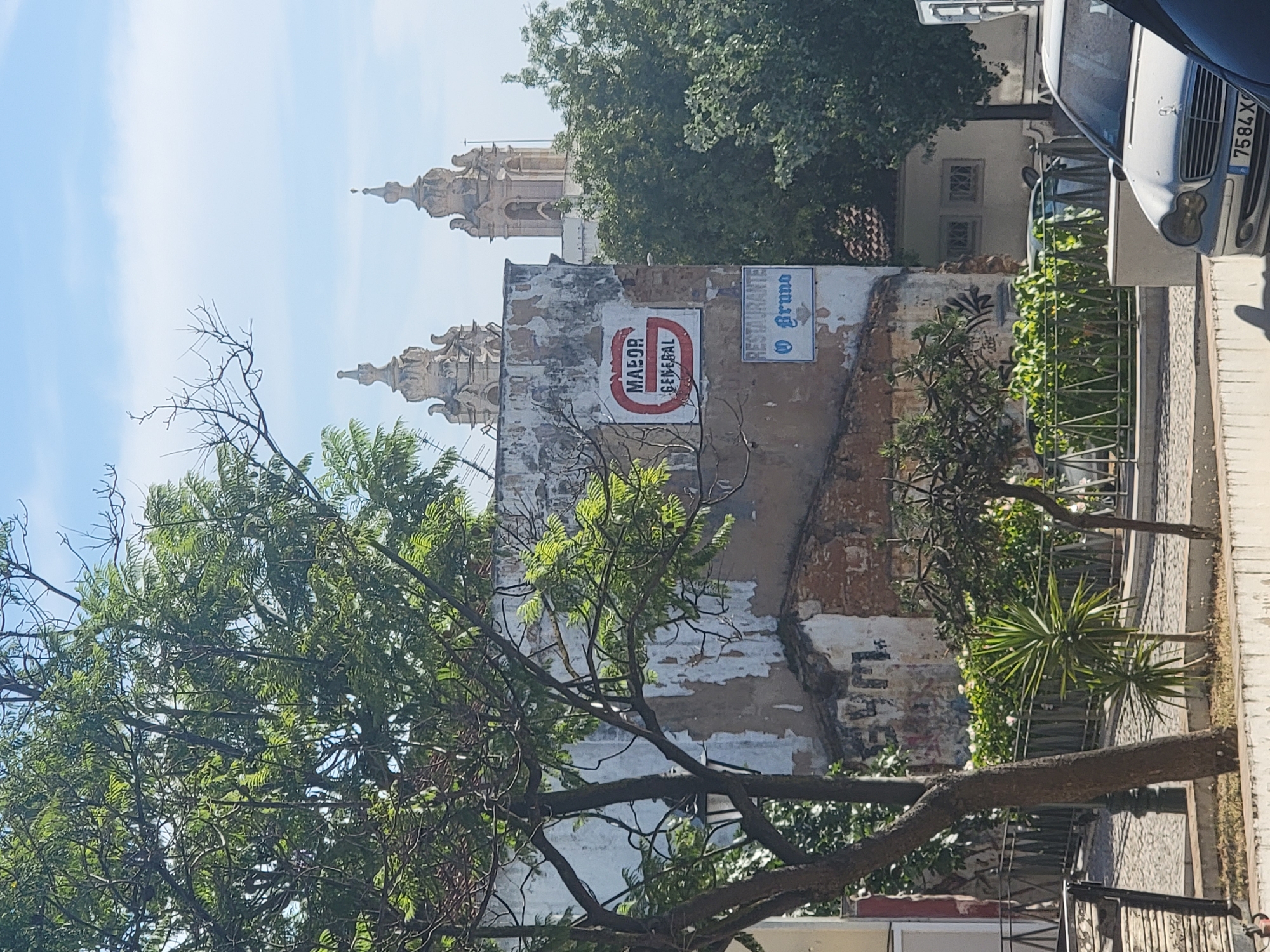 The image captures a street scene in Faro, Portugal, under a clear blue sky with scattered clouds. In the foreground, a large tree with dense green foliage dominates the left side of the frame, its branches casting shadows. Behind the tree, a weathered building stands, its facade showing signs of decay with peeling paint and exposed brickwork. Two signs are affixed to this building: a larger, circular red and white sign with the text "MAJOR GENERAL" and a smaller, rectangular blue and white sign that reads "RESTAURANTE O Bruno".
Further in the background, two ornate church towers with distinctive architectural details rise above the trees, suggesting a significant religious building nearby. A lush green hedge lines a pathway in the middle ground, and beyond that, a silver Mercedes-Benz car with the license plate "7584 X..." is visible, parked on the street. The ground appears to be paved with cobblestones in some areas. The overall impression is of an urban environment where old architecture meets newer elements, with a touch of greenery and a hint of daily life.