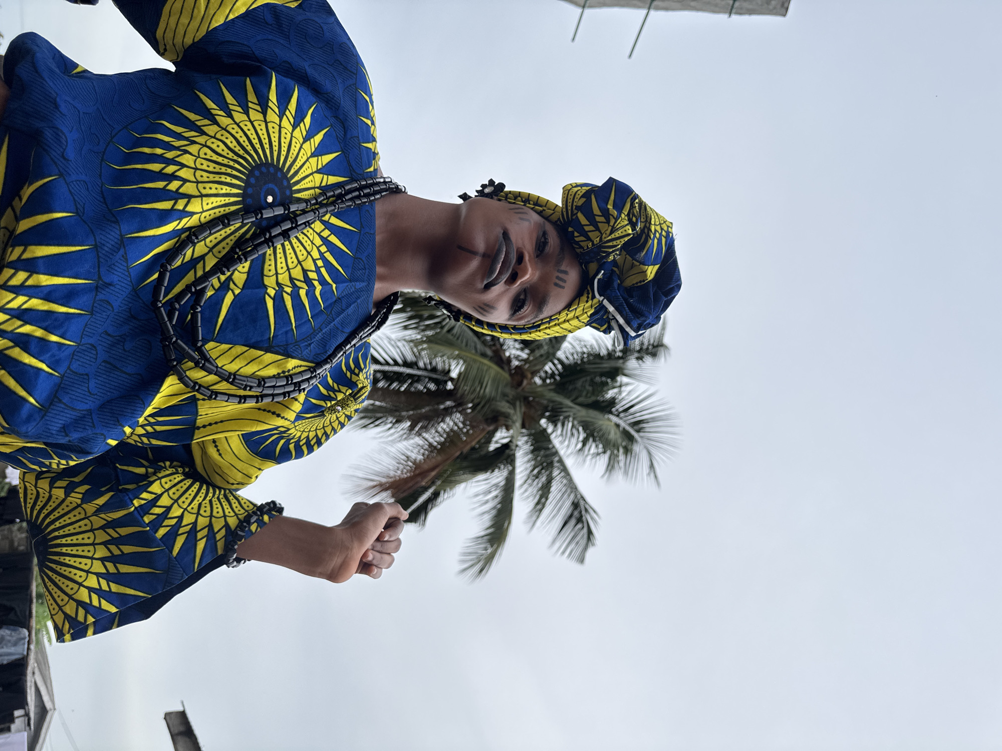 A medium shot shows a woman in traditional Nigerian dress standing in Badagry, Nigeria, in front of a palm tree. She is wearing a bright blue and yellow print tunic and matching head wrap. Her face is painted with tribal markings. A strand of black beads wraps around her neck. She is grasping a palm frond in her left hand. The sky is overcast.