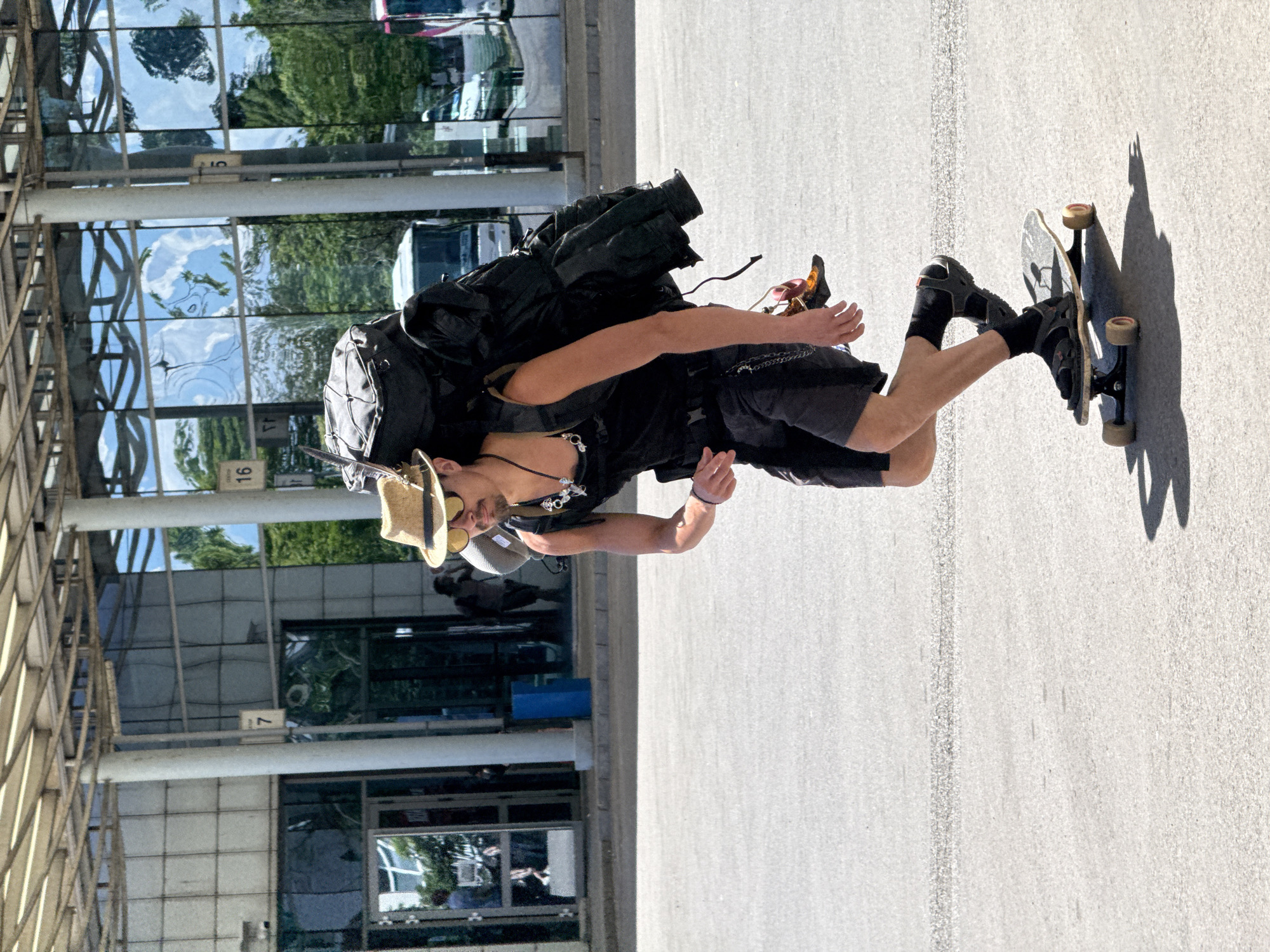 A young man with a straw hat and sunglasses is skateboarding with a large backpack on his back. He is wearing a black tank top, black shorts with a chain accessory, and black sandals with socks. He is carrying an extra pair of shoes attached to his backpack. The background shows a modern building with glass windows reflecting the sky and trees, and what appears to be a bus station with buses visible. The scene is outdoors on a paved surface, likely a street or plaza. The image is taken from a medium distance, showing the man and his skateboard in the foreground and the building and buses in the background. The lighting suggests it is a bright, sunny day.