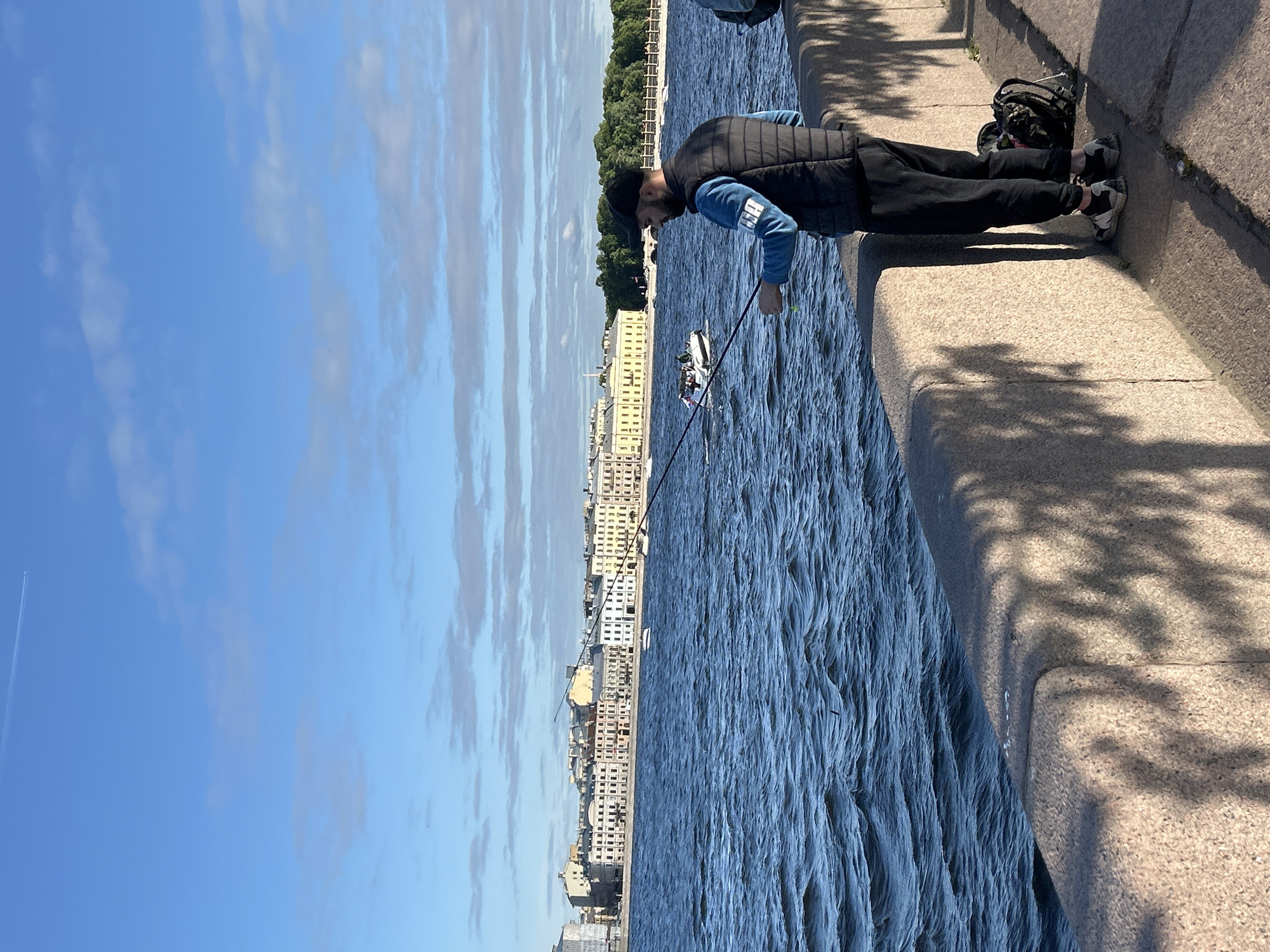 A man wearing a black vest, blue long-sleeved shirt, black pants, and a black baseball cap stands on a stone embankment, fishing with two rods. A black backpack rests on the ground next to him. The man is facing away from the camera, looking out at a wide, choppy blue body of water. Across the water, a city skyline is visible, featuring a long row of beige and white buildings with numerous windows, characteristic of European architecture. The sky above is a clear blue with scattered, thin white clouds. In the distance on the water, several boats are present, including a white motorboat with passengers. The embankment wall is made of large, light-colored stone blocks, with shadows of foliage cast upon its surface. The sun is shining, creating bright lighting conditions.