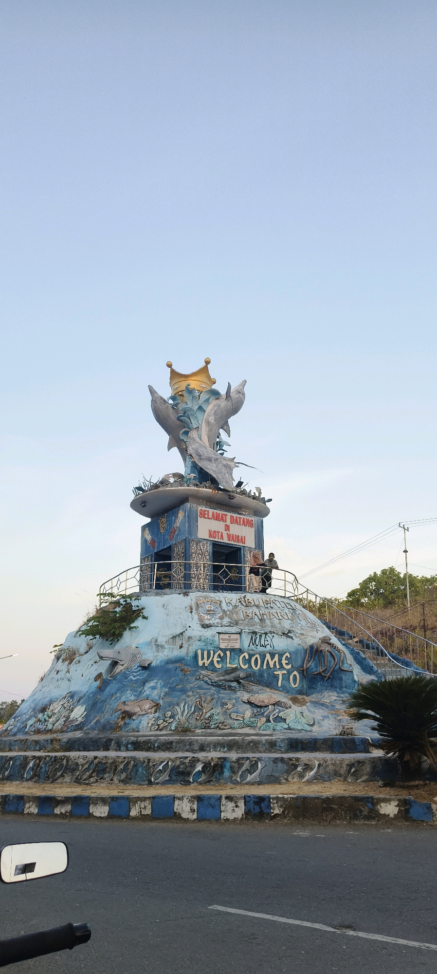 The image shows a monument in the city of Waisai, Indonesia, featuring a sculpture of three dolphins with a golden crown above them. The monument is decorated with marine life motifs, including fish, an octopus, and a shark. Two people are standing on a platform at the base of the monument, overlooking the scene. The monument appears to be made of concrete and painted in shades of blue and white, with some wear and tear visible. The background consists of a clear sky, some trees, and a utility pole with wires. A motorcycle's handlebar and rearview mirror are visible in the foreground, suggesting the photo was taken from a moving vehicle. The monument has signage that reads "SELAMAT DATANG DI KOTA WAISAI" (Welcome to Waisai City) and "WELCOME TO".