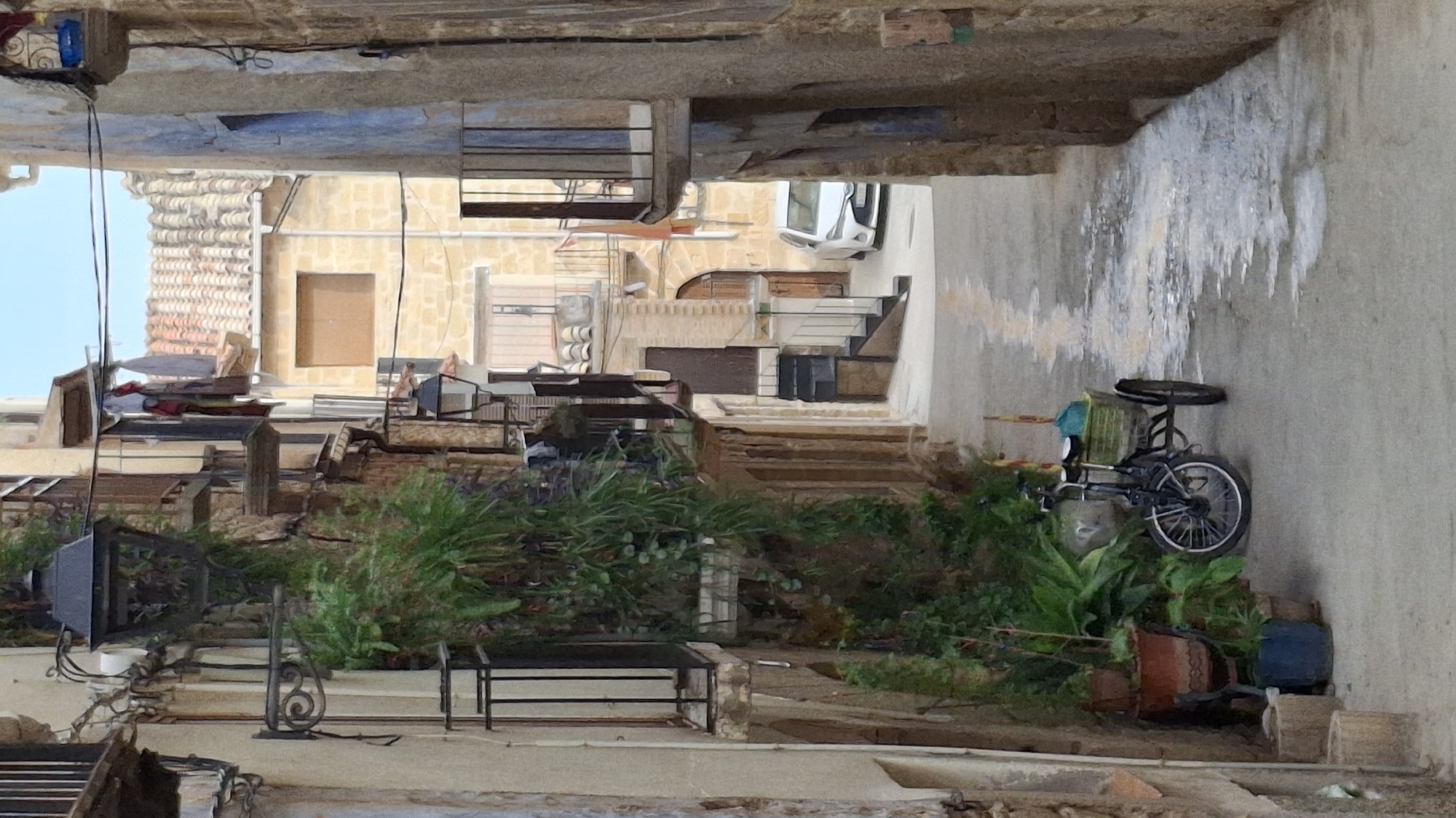 The image depicts a narrow, aged alleyway in the Spanish town of La Fresneda. The buildings on either side are constructed from rough-hewn stone and plaster, with traditional terracotta tiled roofs visible in the distance. Numerous balconies with wrought-iron railings are present, some adorned with lush green plants. Laundry hangs from lines strung between buildings, suggesting domestic activity. A vintage-style black lantern hangs from the wall on the left.

In the foreground, a three-wheeled bicycle with a basket is parked on the cobblestone pavement. The basket contains a blue plastic bag and some other items. A cluster of potted plants is situated to the left of the bicycle, including a large terracotta pot with green foliage. The pavement is damp and shows reflective patches, indicating recent rain or water spill. Further down the alley, a white car is parked. The overall atmosphere is one of an old, perhaps slightly neglected, Mediterranean town.