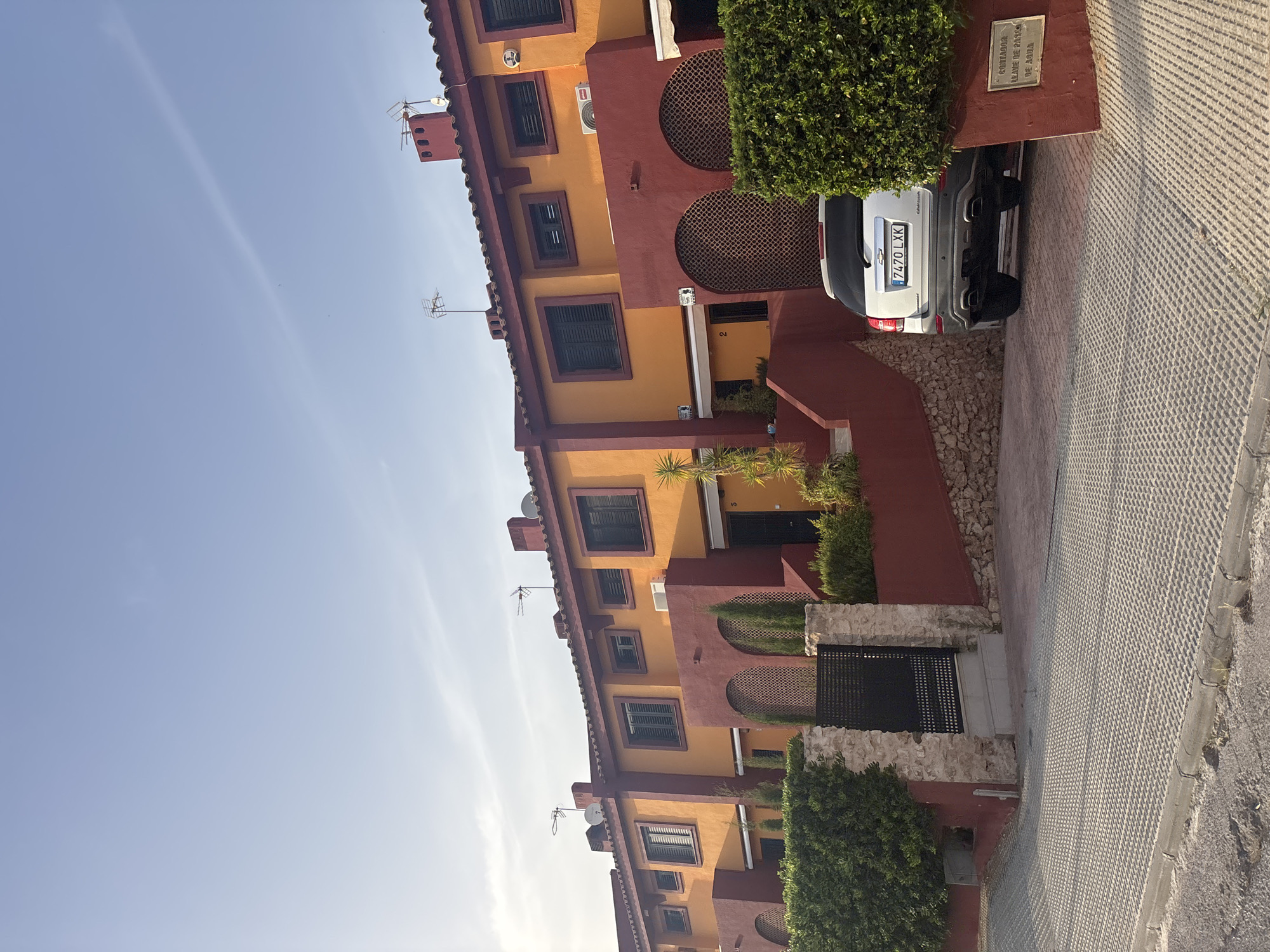 The image captures a row of attached houses in Urbanización Cabo Roig, Spain. The houses are painted in bright orange and deep red colors, with tiled roofs. Each house has dark-shuttered windows. In front of the houses, there are well-maintained green hedges and stone walls. A black metal gate is visible on the left side of the image. A silver Chevrolet Captiva SUV is parked in the driveway of one of the houses, facing away from the camera. The license plate reads "7470 LXK". The ground in front of the houses and driveway is paved with interlocking concrete blocks. The sky is a clear light blue with a faint contrail visible. A metal plaque with Spanish text is mounted on a red wall to the right of the car.