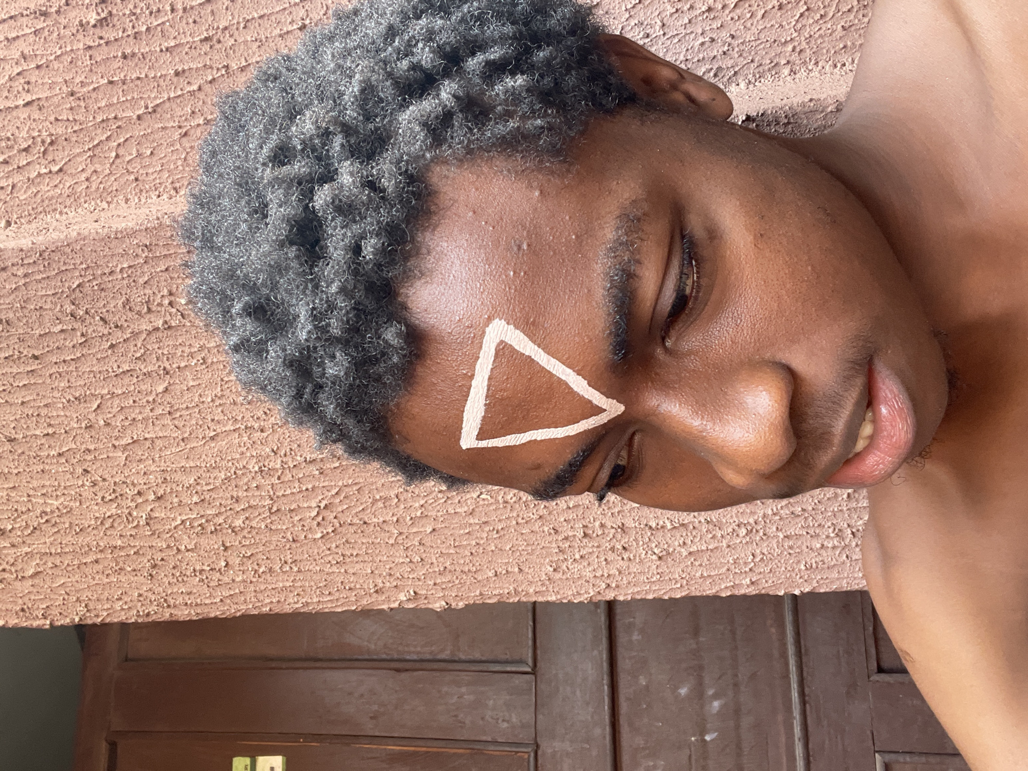 A medium close-up, eye-level shot captures a young man with dark skin and tightly curled, short dark hair, lying down with his head and shoulders visible. Painted on his forehead is a white triangle. He is partially facing the camera, with his gaze directed downwards. The background consists of a textured, light brown wall above and a dark wood surface with door paneling below. The location is plausibly Nkpor, Nigeria, based on the subject's likely ethnicity and potentially the architectural style of the wooden door panels.