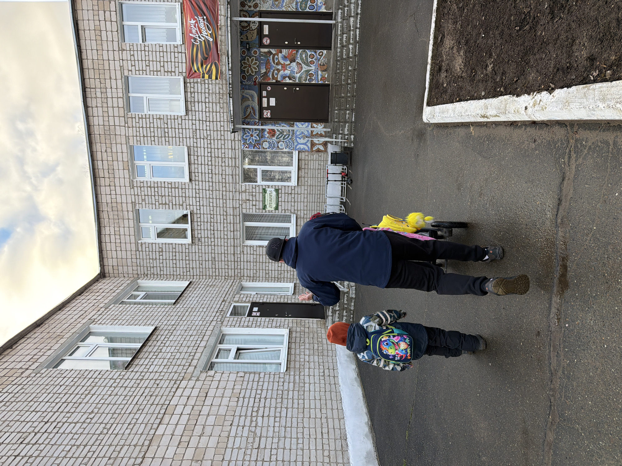 The image captures an outdoor scene featuring a multi-story building and two individuals. An adult, dressed in a dark blue jacket, black pants, and a dark baseball cap, is seen from behind, pushing a small cart or stroller with yellow items. Walking slightly ahead and to their right is a child, also viewed from the back, wearing a dark jacket, an orange beanie, and a brightly colored backpack adorned with cartoon-like figures. They are walking on a dark, damp asphalt path alongside the building.
The building's facade is constructed from light-colored, patterned bricks or tiles, featuring numerous white-framed windows. A prominent main entrance is intricately decorated with colorful mosaic patterns in shades of blue, brown, and white, surrounding two dark brown doors. Above one section of the building, a large red and orange banner hangs, displaying a ribbon motif. Further along the building's side, another entrance is visible, with a green sign above it. To the right of the paved path, there is a raised planter bed filled with dark soil, edged with white concrete. The sky is overcast.
Visible text includes "С ДНЕМ ПОБЕДЫ!" (Happy Victory Day!) on the large banner, and "ЗУБОТЕХНИЧЕСКАЯ ЛАБОРАТОРИЯ" (Dental Laboratory) on the green sign above the second entrance. Small, indistinct signs are also present on the dark brown doors of the main entrance. The overall setting appears to be a residential or public area in Chaikovsky, Russia, during daylight hours.