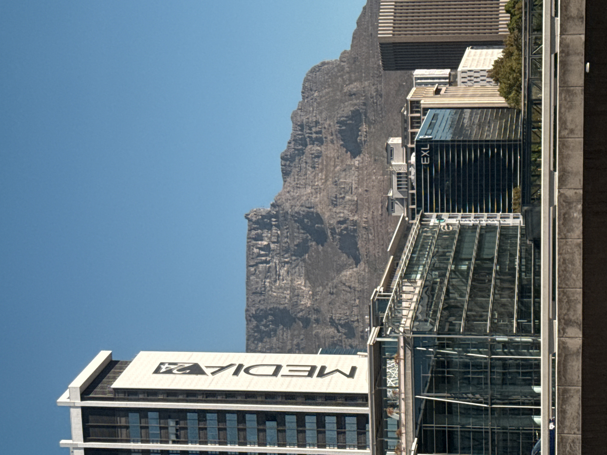 The image captures the urban landscape of Cape Town, South Africa, with Table Mountain prominently featured in the background. In the foreground are modern high-rise buildings with glass facades, reflecting the cityscape. One building prominently displays the "MEDIA 24" logo, while another has the "EXL" name on it. The sky is clear and blue. The image is taken from an elevated perspective, showcasing the juxtaposition of the natural mountain landscape and the urban development of the city.