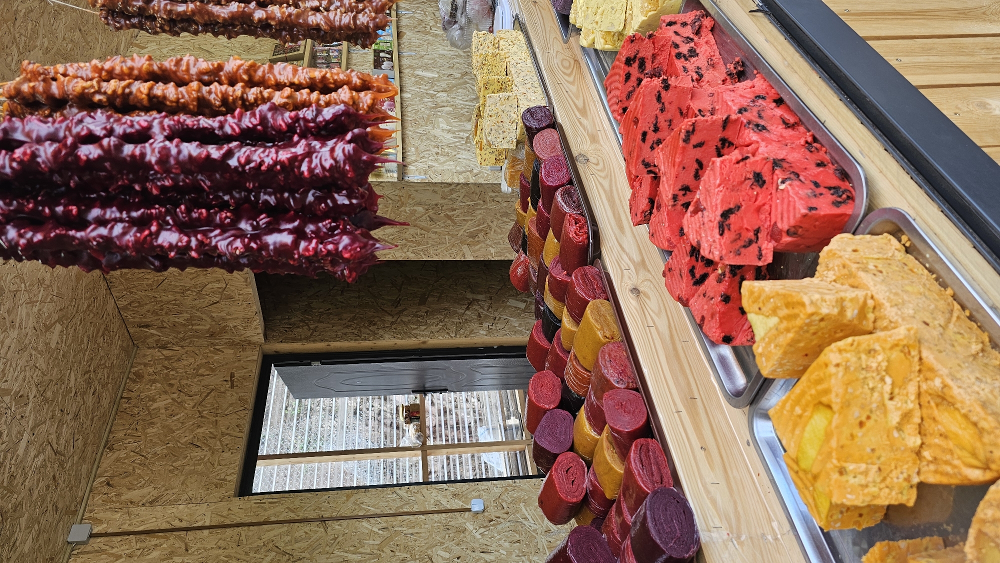 The image depicts a market stall filled with various types of dried fruit leather and candied fruit products. In the foreground, several trays display brightly colored, rectangular blocks of what appear to be fruit leather or a similar confectionery. One tray holds orange-colored blocks, some with visible fruit pieces, while another tray contains vibrant red blocks studded with dark fruits. Behind these, a long row of rolled fruit leathers in various colors—purple, red, yellow, and orange—are neatly arranged on a wooden surface. Further in the background, more stacked blocks of light yellow, possibly nut-filled, confectionery are visible. Hanging from the ceiling are long strands of dark red and orange dried fruit products, resembling churchkhela. The stall is constructed with wood-paneled walls and ceiling, and a wooden counter displays the goods. An open doorway leads to an area with a window, through which some shelves with jars can be seen. The location is identified as Красный Курган, Russia. There are no people visible in the image.