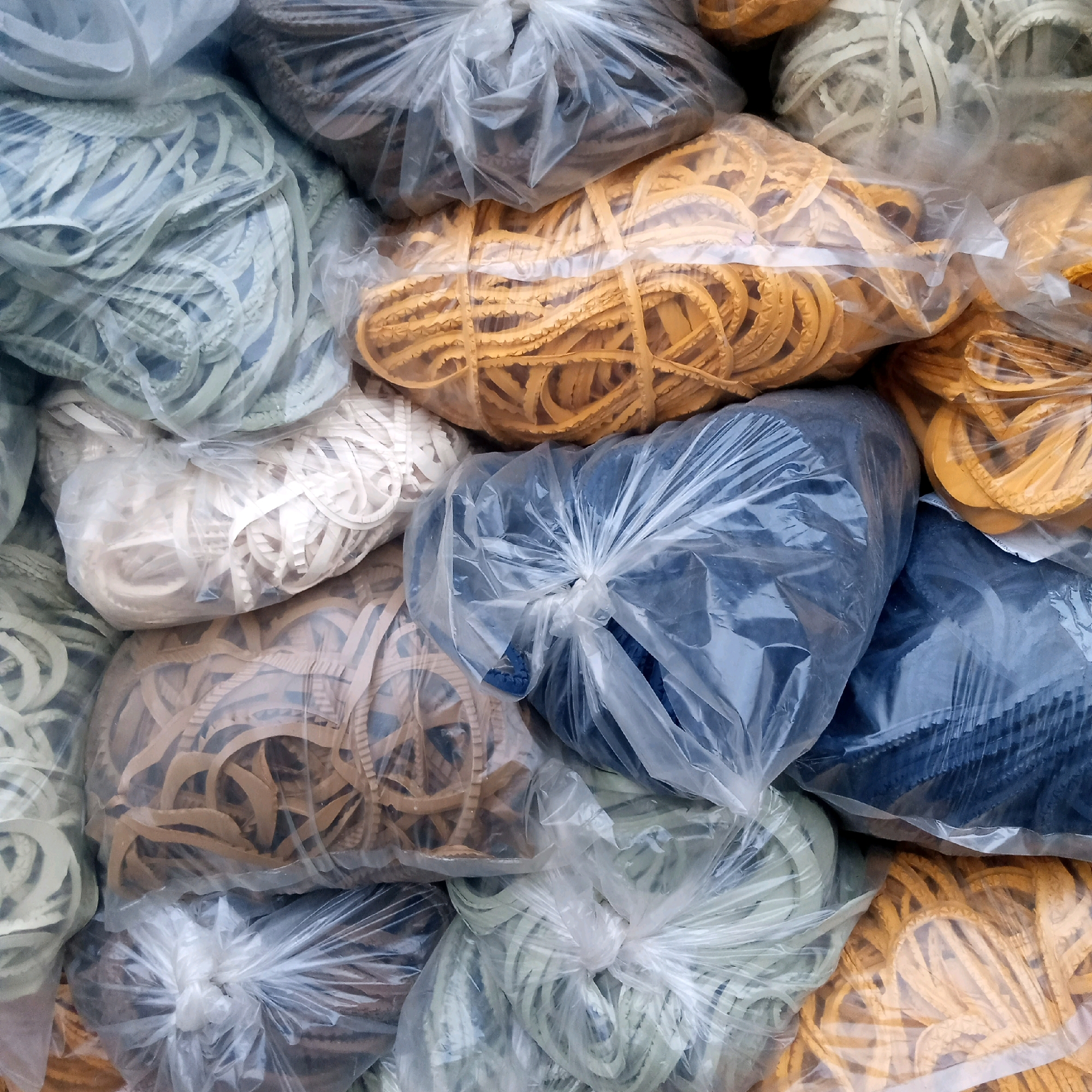 This is a close-up, eye-level photograph of a large pile of plastic bags filled with various colored rubber bands. The rubber bands are coiled within the bags, and some of the bags are tied shut with white twist ties. The colors of the rubber bands include light blue, tan, yellow, and dark blue. The overall impression is one of bulk storage or retail packaging. The scene is undated and does not provide information about the location in Cijeruk, Indonesia. There are no people, actions, or interactions visible in the image. The environment appears to be indoors, possibly a warehouse or a storage facility.