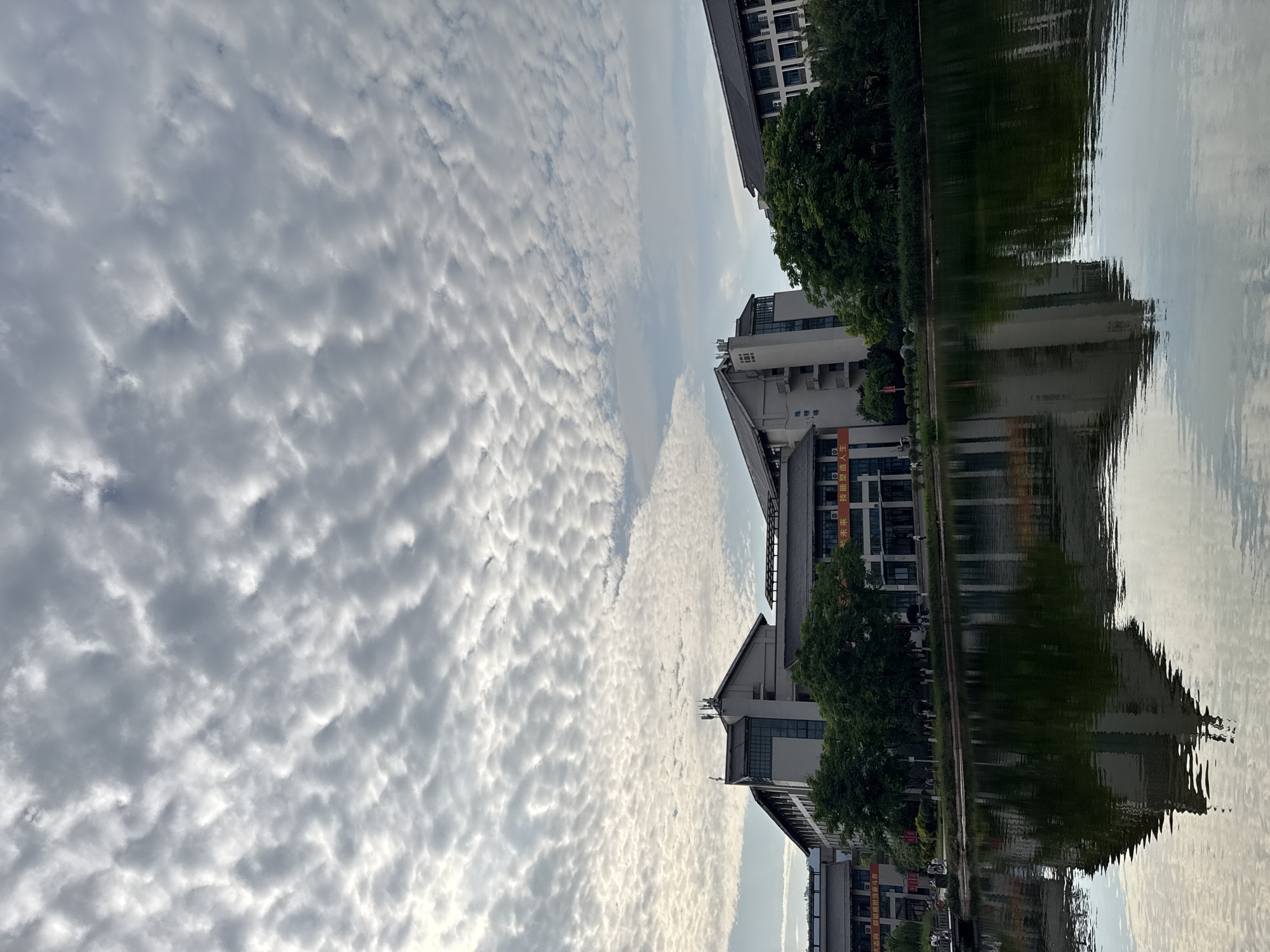 The image shows a landscape with buildings and trees reflected in the water. There are clouds in the sky. The location is likely Jiangjin, China. The scene includes buildings, trees, water reflection, and clouds. There are no discernible people, actions, or interactions.
