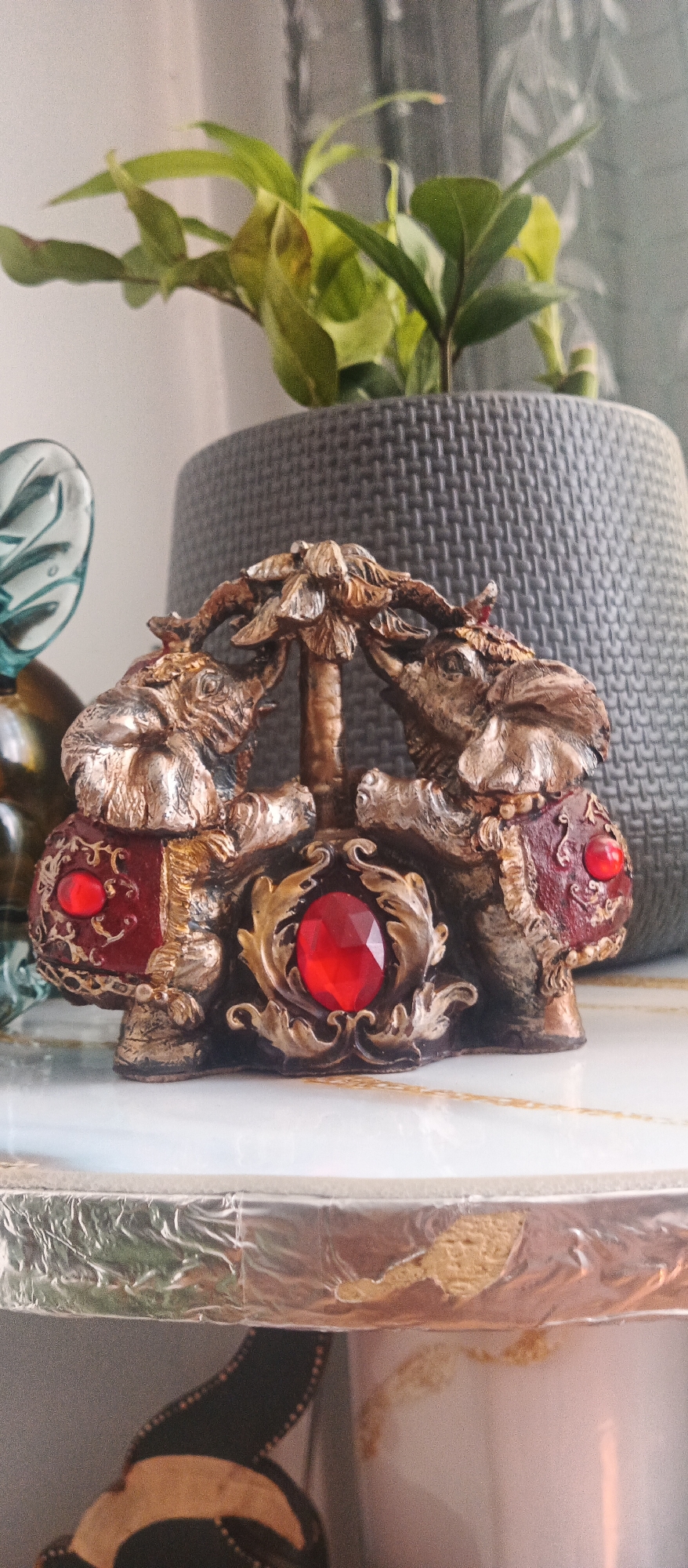 A close-up, eye-level, medium shot captures a decorative, metallic figurine of two elephants facing each other with their trunks raised and touching in the center, creating an arch. The figurine is intricately detailed with a golden-bronze finish, appearing antique or weathered. The elephants' bodies are adorned with red, gem-like embellishments on their sides and a larger, faceted red jewel is prominently displayed at the base of the figurine, framed by ornate golden scrolls. The figurine rests on a white surface with a marble-like pattern and a subtle golden inlay. Behind the figurine, a textured, dark grey planter holds a lush green plant with broad leaves, its foliage partially obscuring the upper portion of the planter. The background is softly blurred, suggesting a domestic indoor setting.