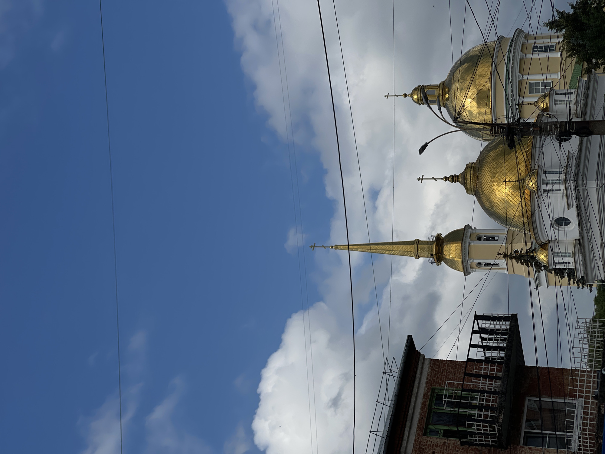 The image shows the golden domes and spire of the Saint Michael's Cathedral in Izhevsk, Russia, against a backdrop of a bright blue sky with scattered clouds. In the foreground, a brick building with a balcony is partially visible on the left, and electrical wires crisscross the scene, obscuring parts of the cathedral. The cathedral's ornate architecture, including its multiple domes topped with crosses, is clearly depicted. The overall impression is of an urban landscape dominated by religious architecture, with the presence of everyday infrastructure like buildings and wires.