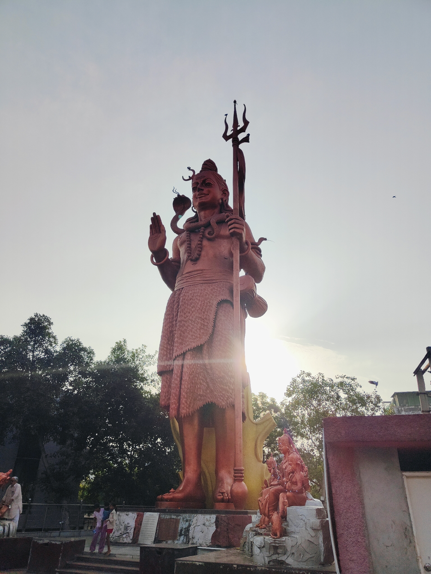 A low-angle, wide shot captures a massive, reddish-brown statue of Lord Shiva standing tall against a bright, hazy sky in Gurgaon, India. The statue is elaborately detailed, with Shiva depicted holding a trident in his right hand and raising his left hand in a gesture of blessing. A serpent is coiled around his neck. To the right of the main statue, smaller statues of Shiva and other deities are visible, seated on a rocky base. Trees with dense green foliage frame the background, and a building with a reddish-brown roof is present on the far right. Several people are present in the lower part of the frame, including a man in white attire on the left, and two children in colorful clothing standing on steps in the center. The sun is low in the sky, casting a bright lens flare across the image, suggesting it is either sunrise or sunset. The overall impression is one of grandeur and spiritual significance.