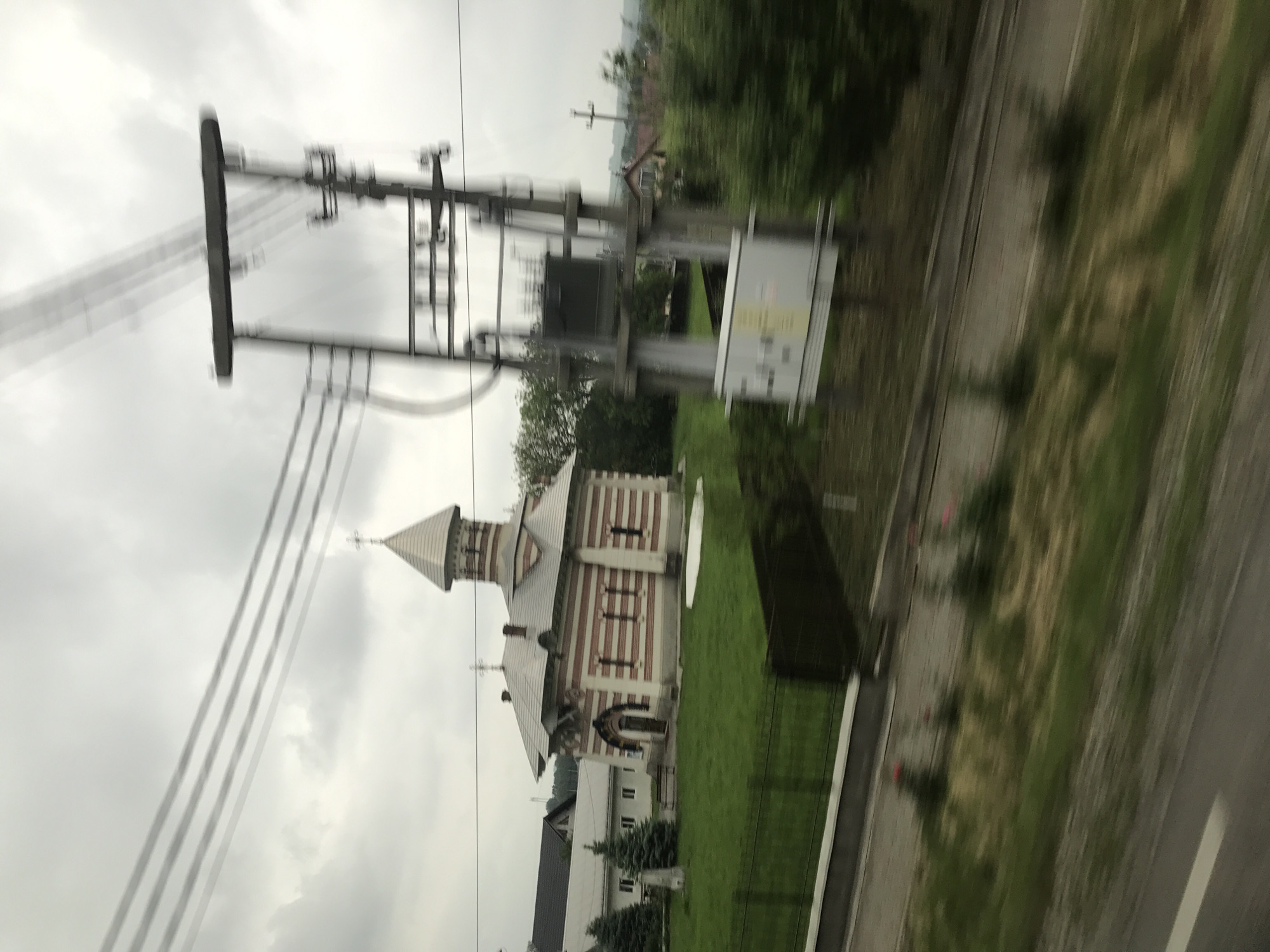 The image displays a motion-blurred landscape in Drăgușeni, Romania, captured from a moving vehicle under an overcast sky. The central subject is the Monastery of Drăgușeni church, identifiable by its distinct architecture featuring a facade with horizontal red and light-colored stripes, a grey conical spire, and prominent crosses affixed to the spire and the main roofline. Behind the church, a plainer white building with a dark roof is visible, nestled within a manicured green lawn. Several evergreen trees are positioned around these structures, with a dark fence separating the lawn from the foreground. Dominating the right and central foreground is a heavily blurred electrical utility pole structure with multiple horizontal wires, indicative of rapid movement. The immediate foreground features a blurred asphalt road with a partial white line and sparse ground vegetation along its edge. Distant residential buildings and additional foliage are visible on the horizon. No individuals are present in the frame.