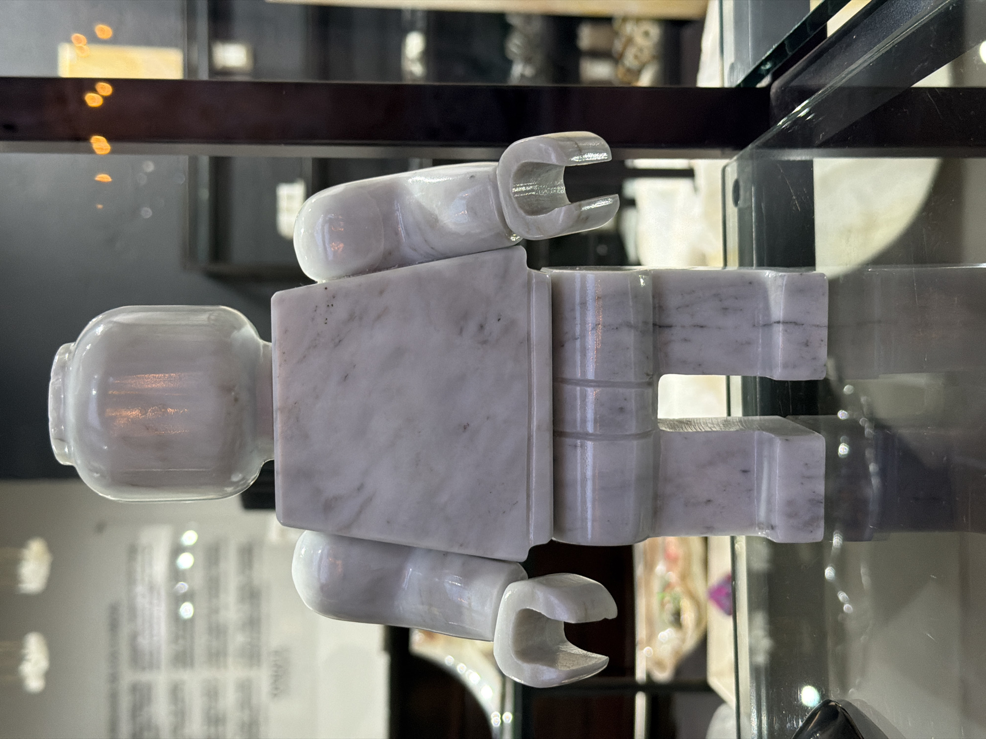 The image features a marble sculpture resembling a Lego figure, displayed inside a glass case. The sculpture is white with grey veining, characteristic of marble. The background is blurred, showing other objects within the case and the environment outside the display, suggesting a retail or museum setting. The location is Domingo Arenas, Mexico.