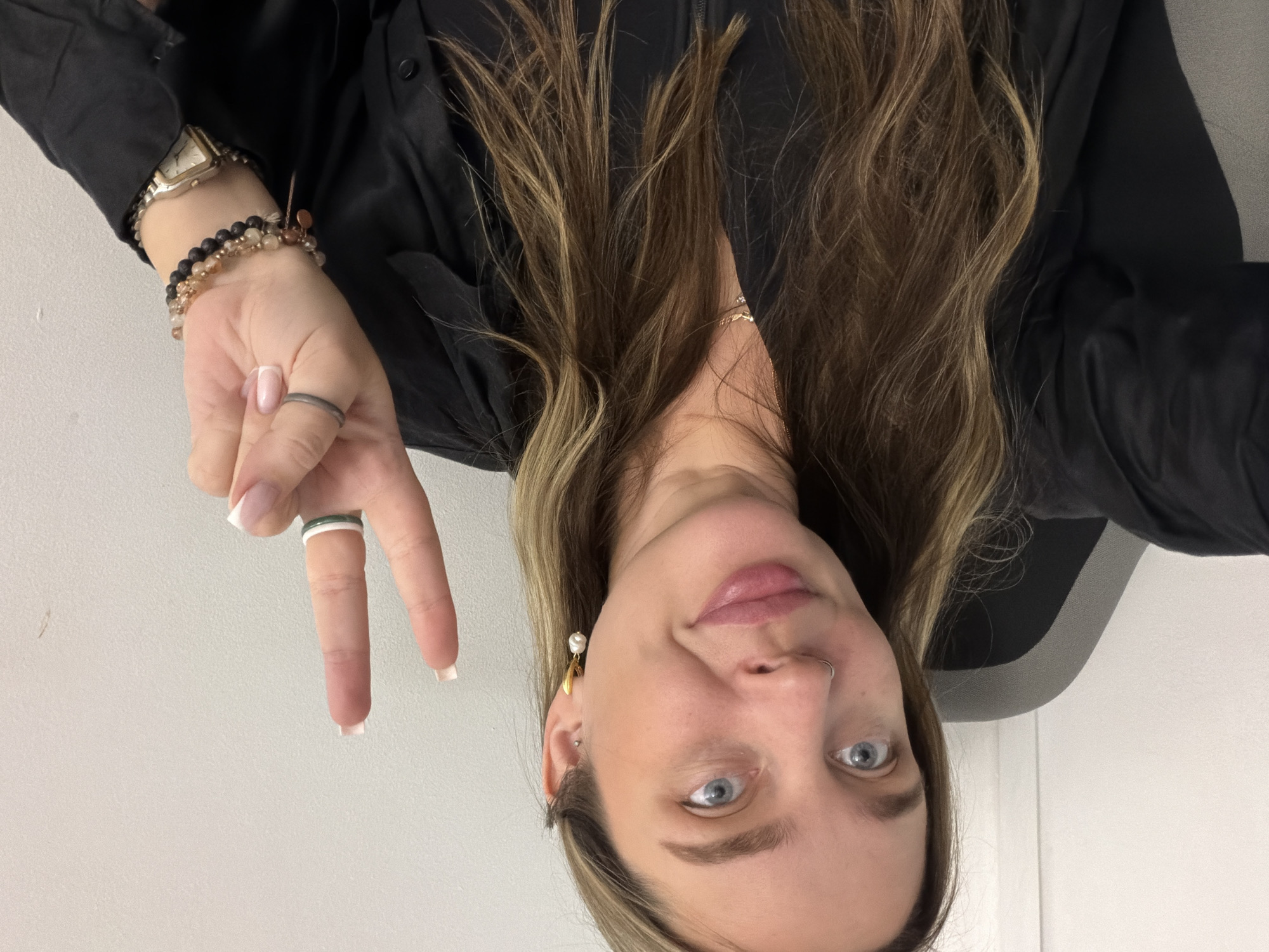 A young woman is captured in a headshot and upper torso selfie, taken indoors against a plain white or light grey background, likely in Moscow, Russia. She has long, light brown hair with blonde highlights, clear blue eyes, and a soft, natural makeup look. She is wearing a black shirt or top. Her left hand is raised to display a peace sign, and she looks directly at the camera with a calm, slightly smiling expression.
On her left wrist, she wears a gold-toned rectangular watch and several beaded bracelets in earthy tones. Her fingers are adorned with multiple rings: a plain grey ring on her middle finger and a white ring along with another thin band on her index finger. Her nails are manicured in a classic French style, featuring light pink bases and white tips. She also has a small silver nose ring on her left nostril, a pearl drop earring on her right ear, and a small stud on her left ear. A delicate gold-toned necklace with a tiny pendant is visible around her neck. No text is visible in the image.