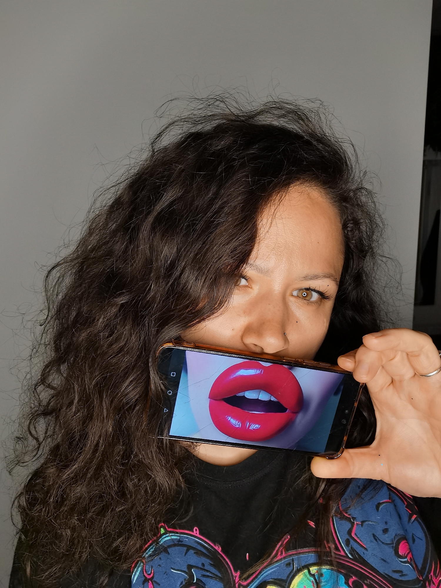 A woman with curly, dark hair is holding a smartphone in front of her mouth. The phone displays an image of bright red lips. She has light skin and brown eyes. She is wearing a black t-shirt with a colorful graphic design. She is standing in an indoor setting with a light gray wall. The location is Chaikovsky, Russia.